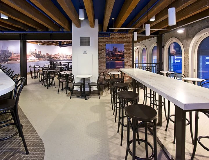 Interior of a modern restaurant or lounge with high tables, bar stools, arched windows, and cityscape artwork on brick wall.