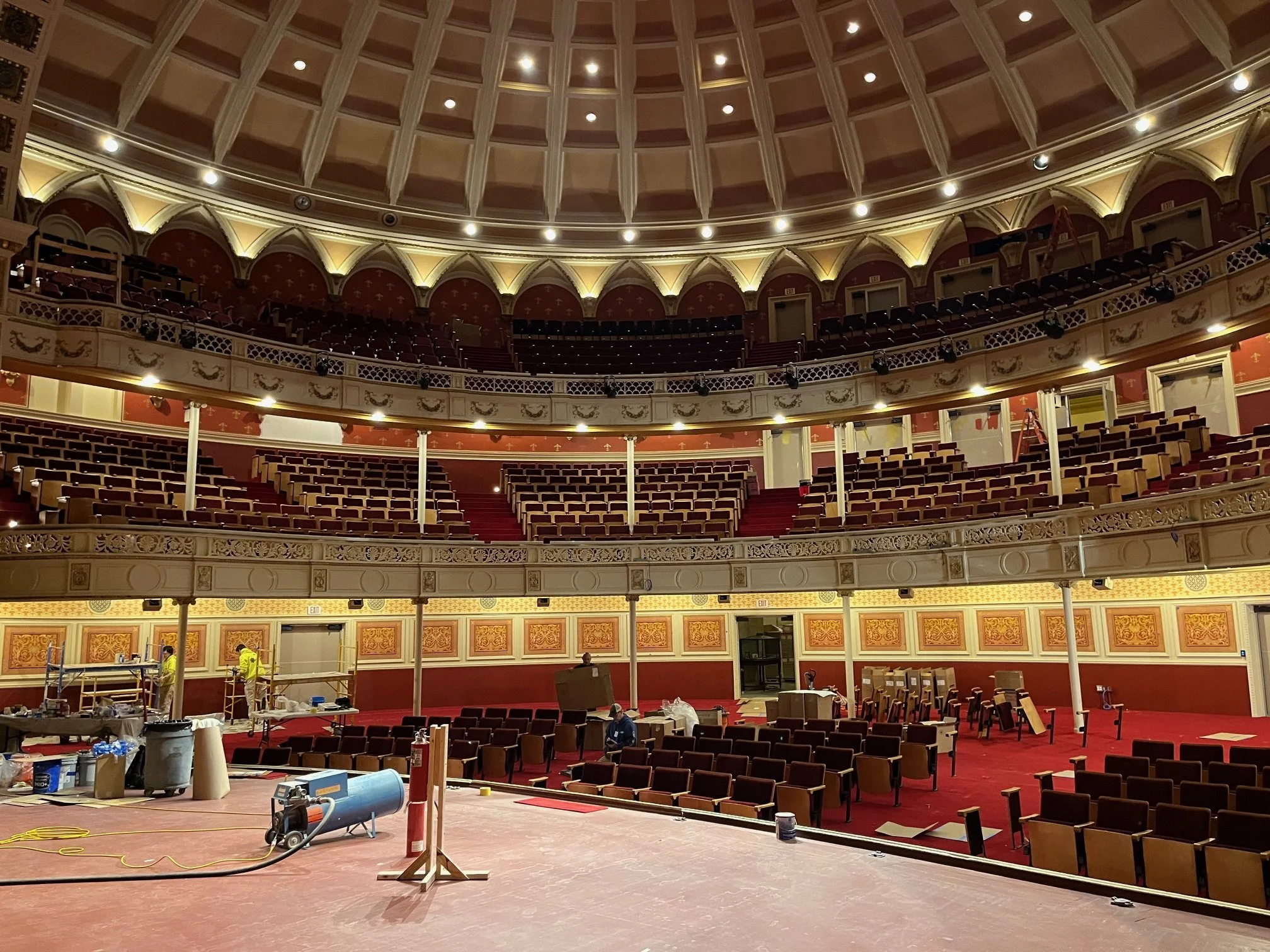 The interior of a theater or auditorium under renovation. The space features multiple tiers of seating, ornate architectural details, and a large, decorated ceiling. Construction workers and equipment are present, indicating ongoing work.