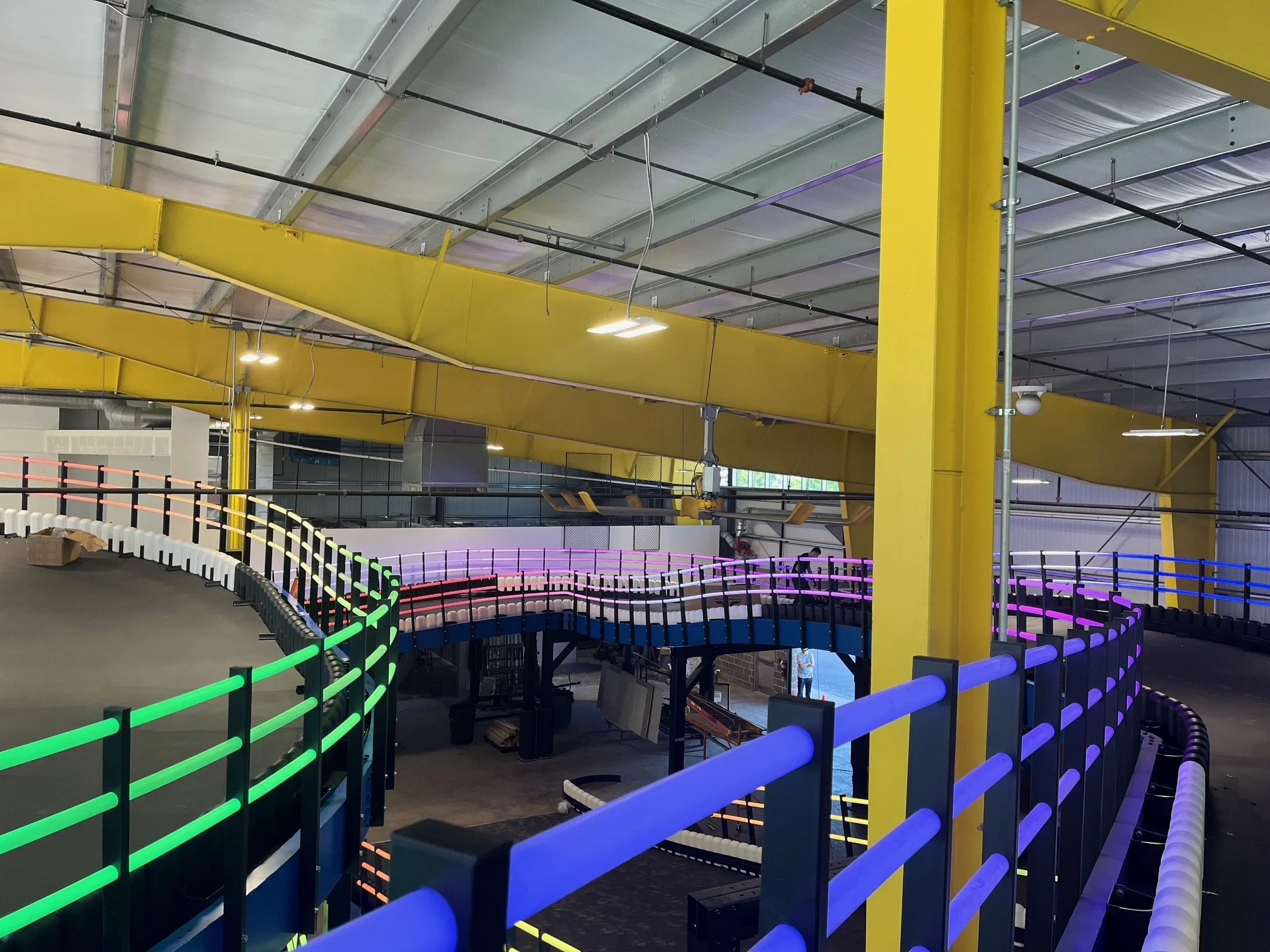 Interior of science center featuring a colorful go-kart track with a vibrant, illuminated safety barrier. The ceiling exhibits industrial design with exposed metal pipes and large yellow structural beams.