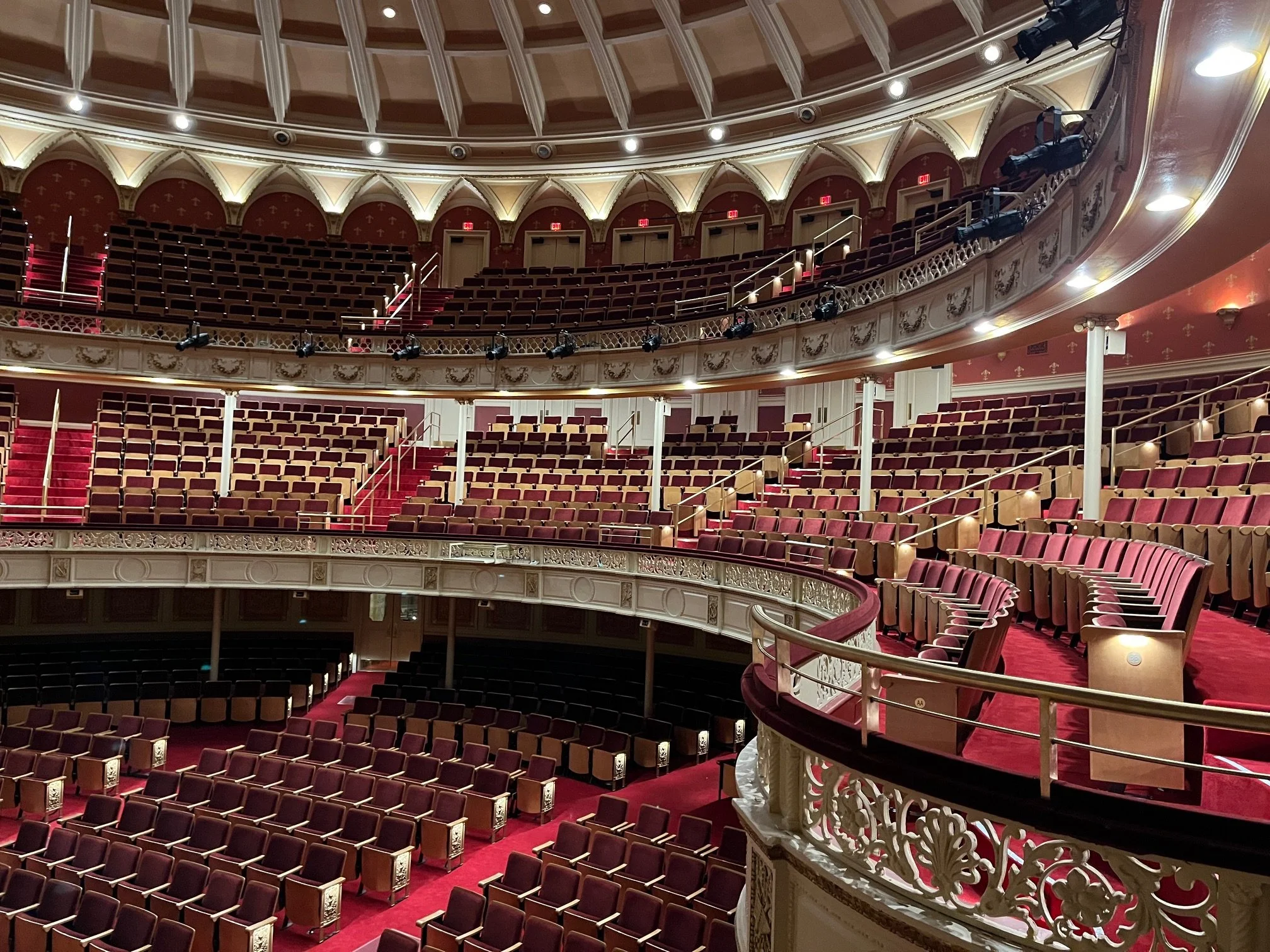 Interior view of an ornate theater with multiple balconies, red velvet seats, and decorative gold and white trims. Stage lighting and curtains are visible at the bottom.