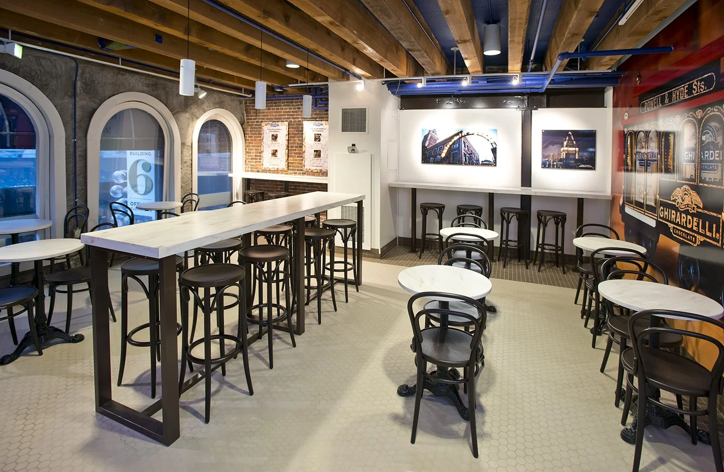 Empty modern cafe with white tables, black chairs, large windows, exposed brick wall, and wall-mounted TVs displaying cityscape images.