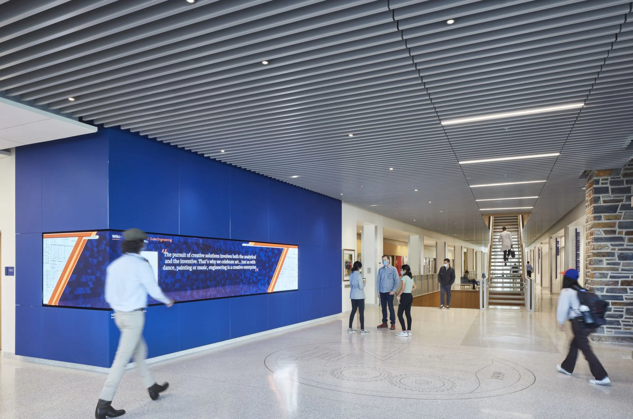 People walking inside a modern building with a blue wall, digital display screen, and escalator in the background.
