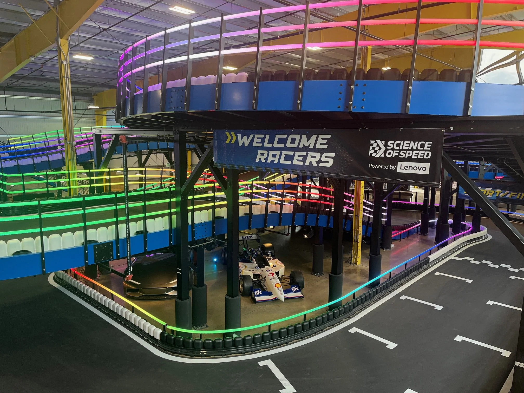 Indoor racing track with a miniature Formula 1 car and a colorful, multi-tiered climbing structure with neon lights, welcome sign, and banners for 'Science of Speed.'