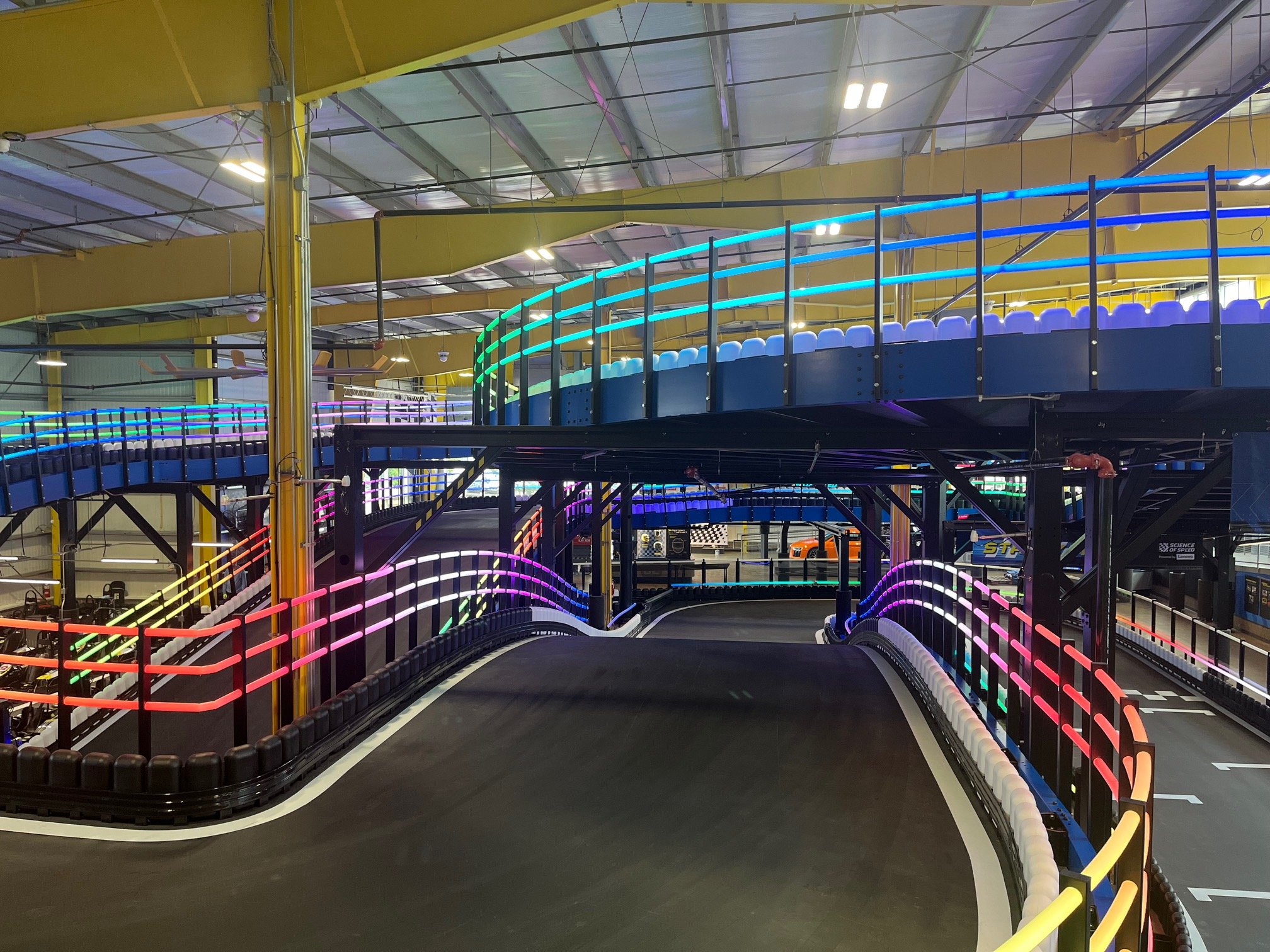 An indoor race track with black flooring, surrounded by illuminated neon-colored railings in pink, orange, yellow, green, blue, and purple hues. The track features sharp turns and is located inside a large industrial-style building with exposed ceili