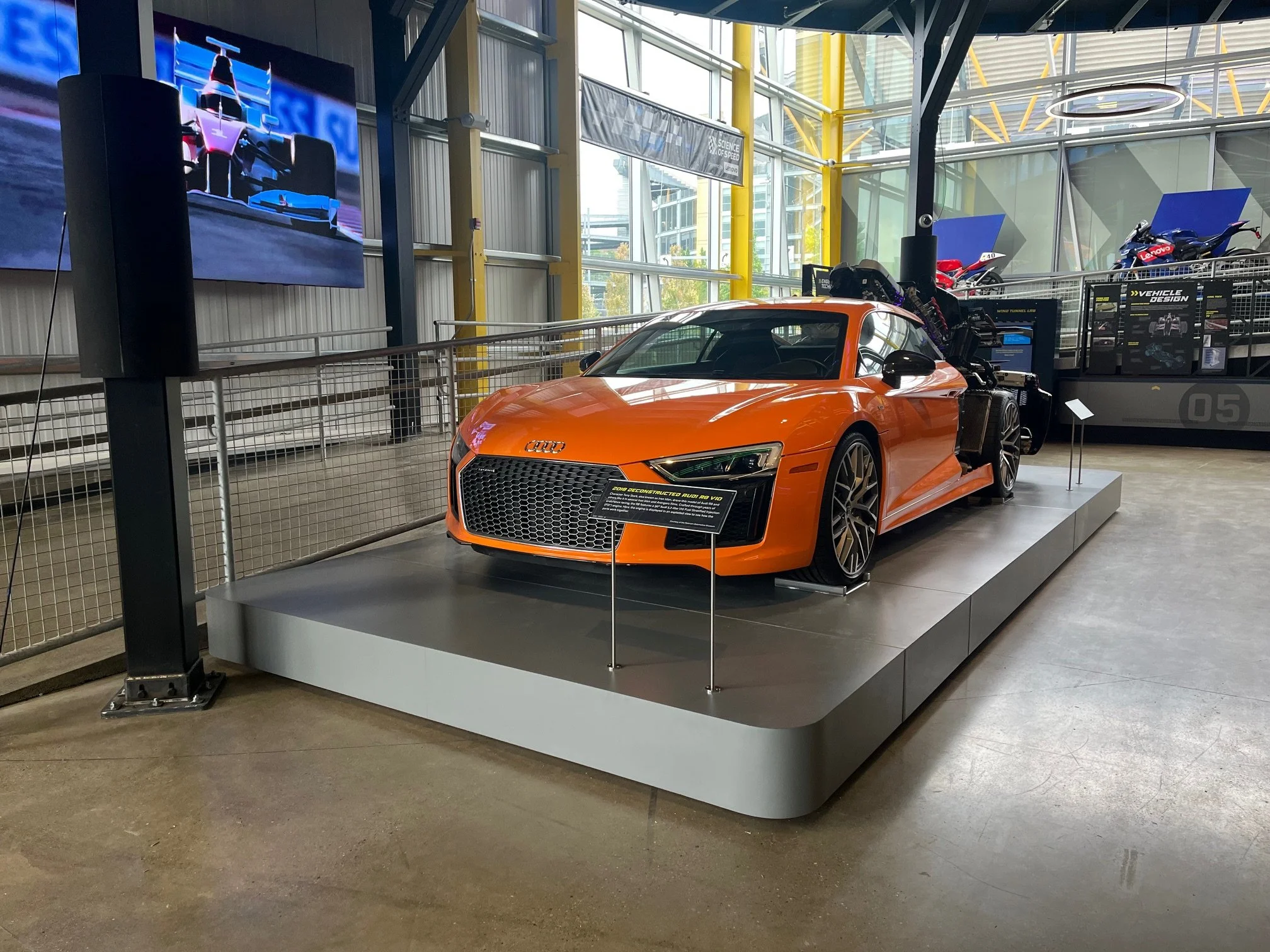 An orange Audi car displayed on a platform at an indoor exhibition or showroom, with motorcycles and display boards in the background.