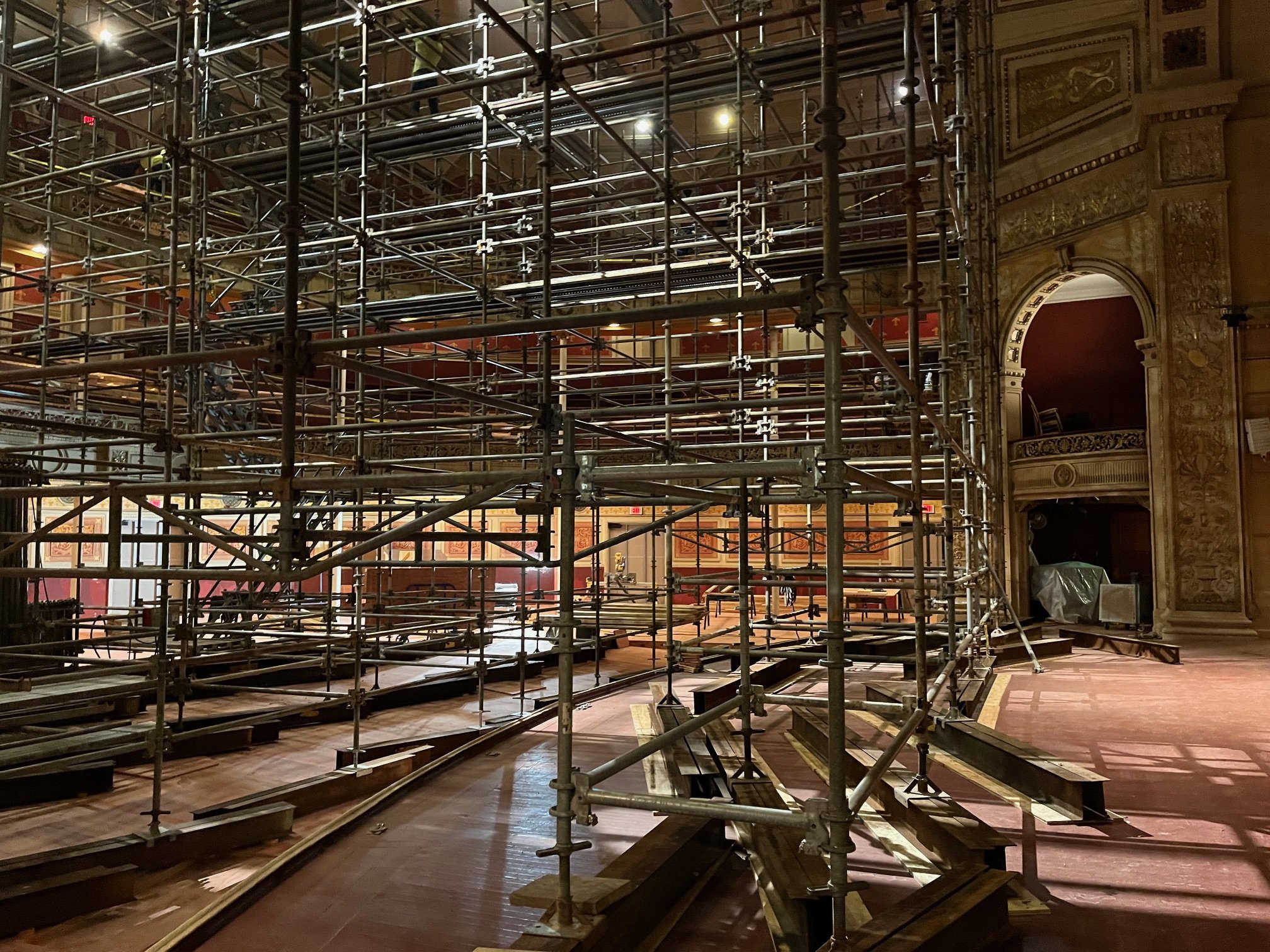 Scaffolding structure set up inside an ornate theater or auditorium, with intricate gold decorations and architectural details visible on the walls and ceiling.