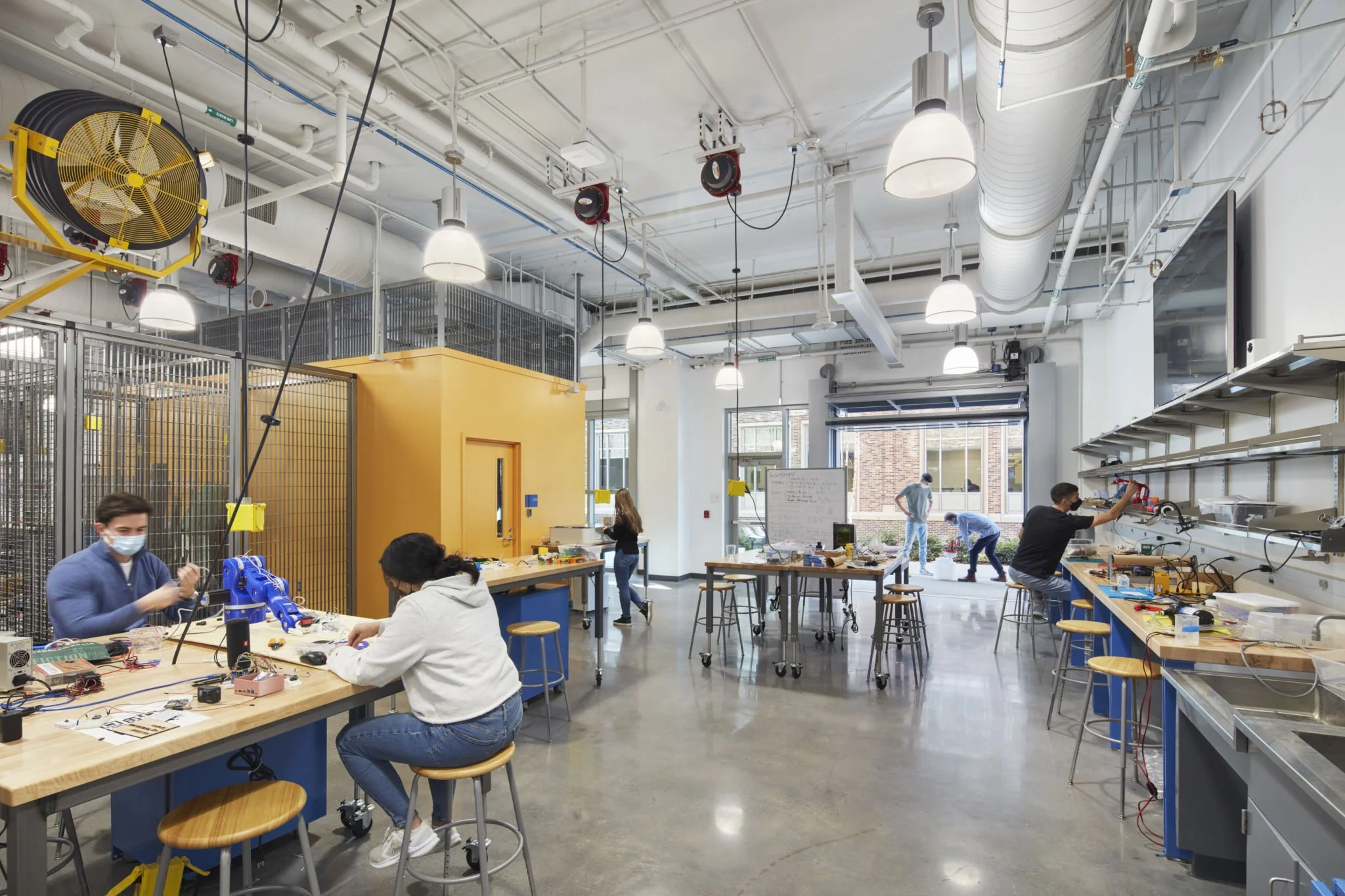 Students working on electronics projects in a large, well-lit classroom or laboratory with high ceilings, white walls, and large windows, featuring workbenches, stools, and various electronic equipment.