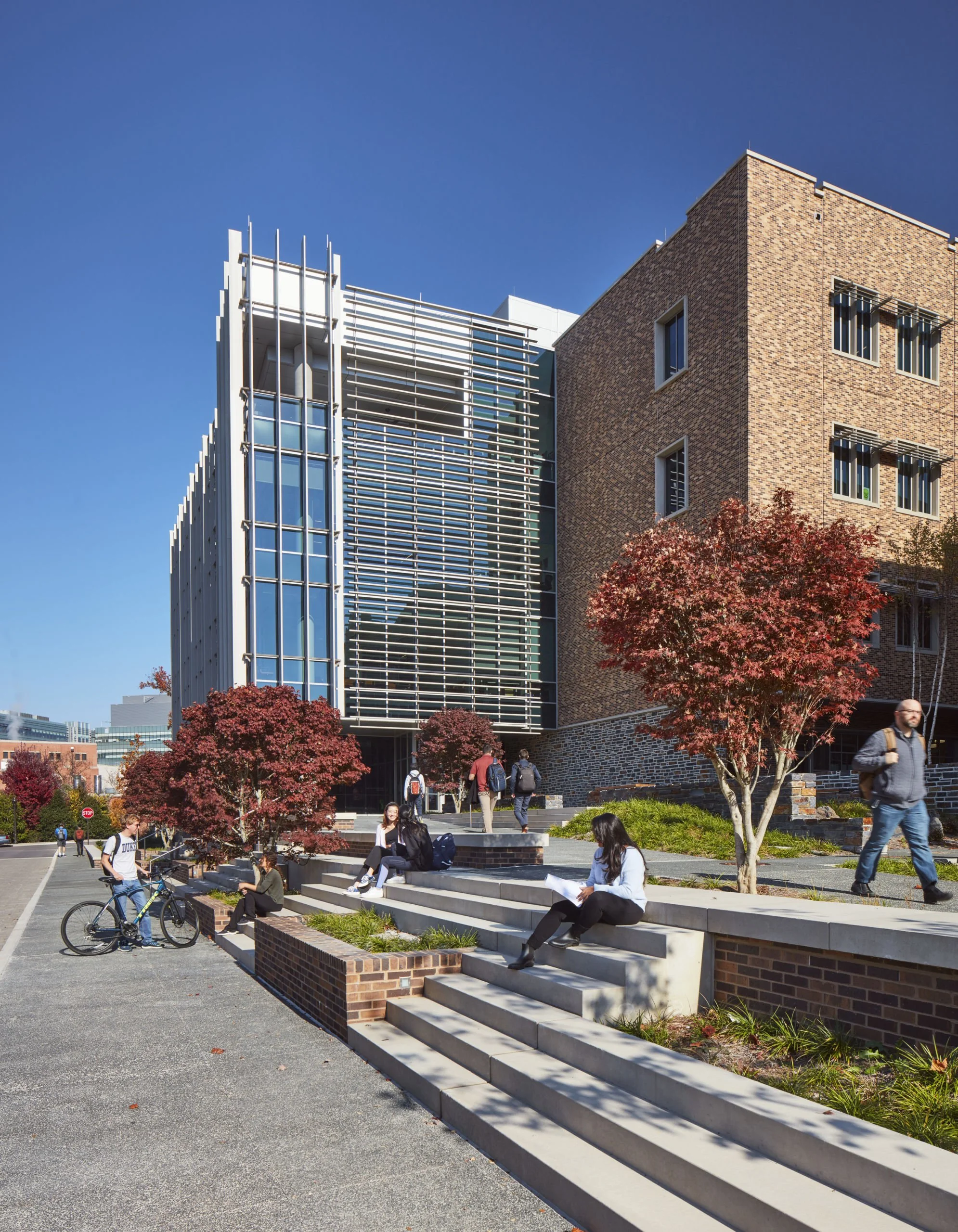 Students relaxing and socializing outside a modern brick and glass university building with trees displaying autumn colors.
