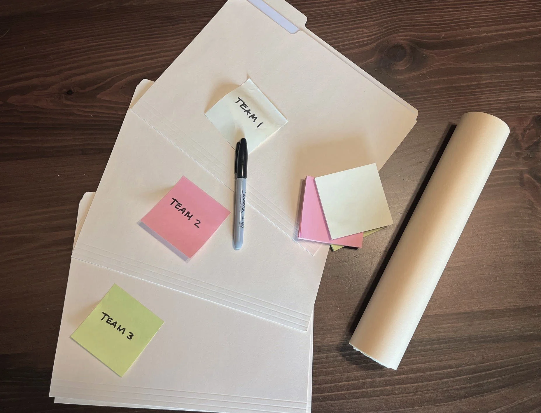 Sheets of paper with sticky notes labeled 'Team 1', 'Team 2', and 'Team 3', a black marker, additional sticky notes, a large sheet of paper, and a roll of brown paper on a wooden table.
