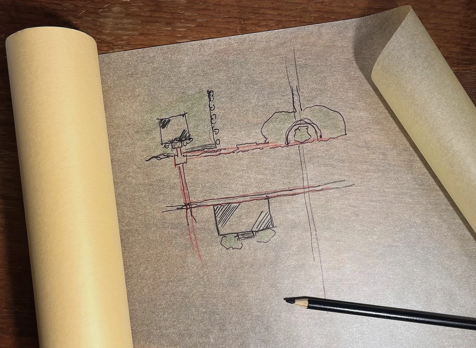 Hand-drawn sketch on yellow tracing paper showing a mechanical or infrastructural diagram with interconnected components, including a large circular element, vertical and horizontal lines, and shaded squares, placed on a wooden surface with a black pen nearby.