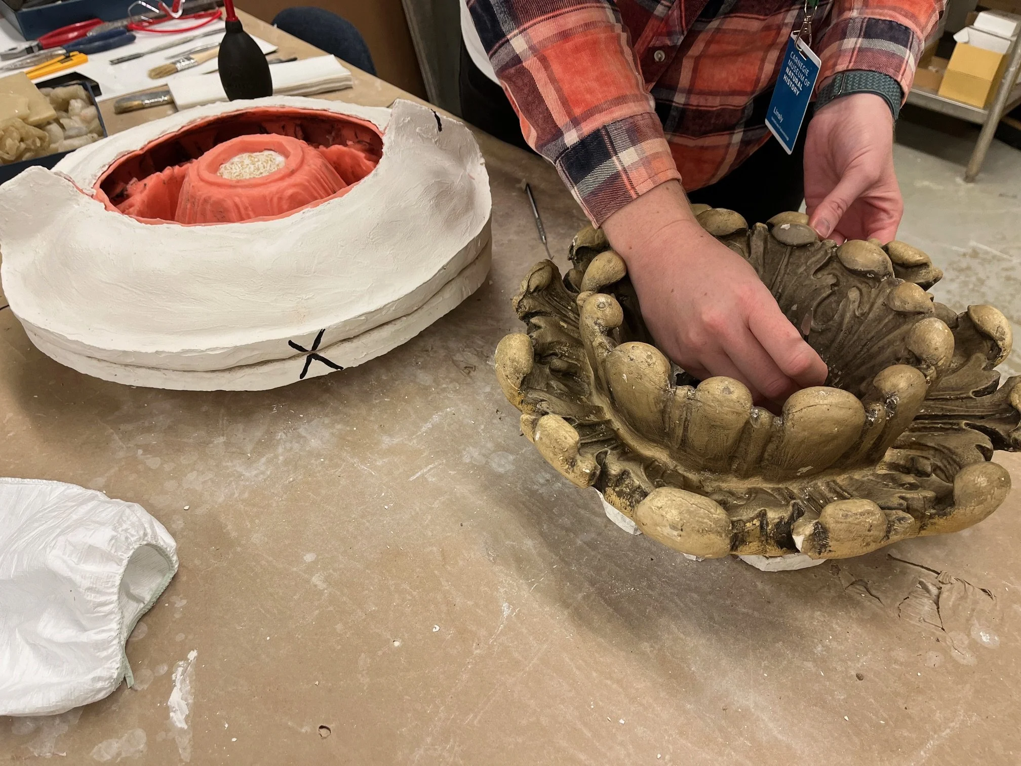 A person wearing a red and black plaid shirt restoring a sculptural element of an historic theater with various tools and materials on the table.