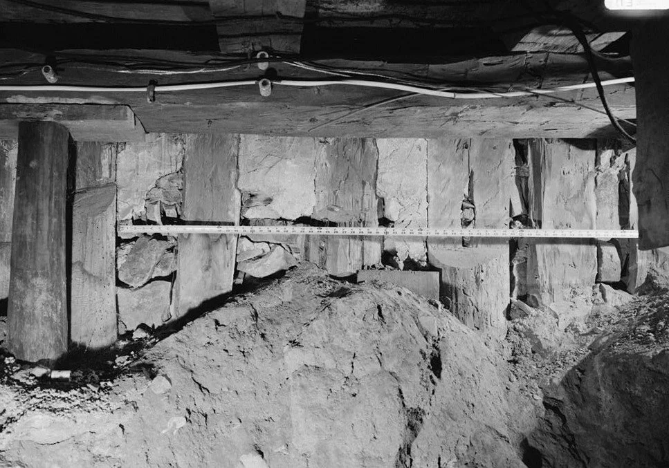 Underground construction site with wooden beams, exposed rocks, a measuring tape, and pipes.