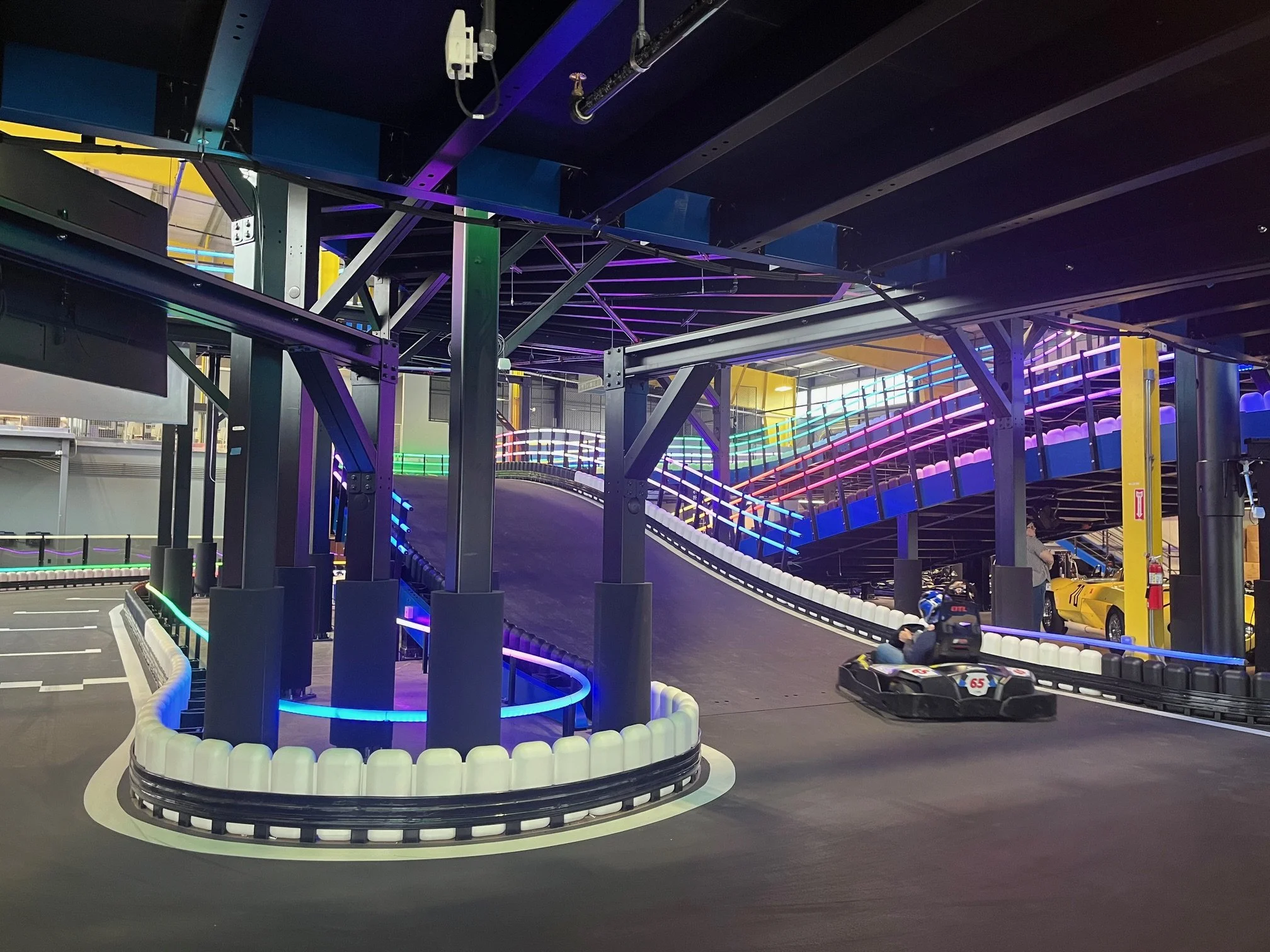 Indoor go-kart track with illuminated, colorful railings and a curved racing lane, featuring a go-kart with a driver.