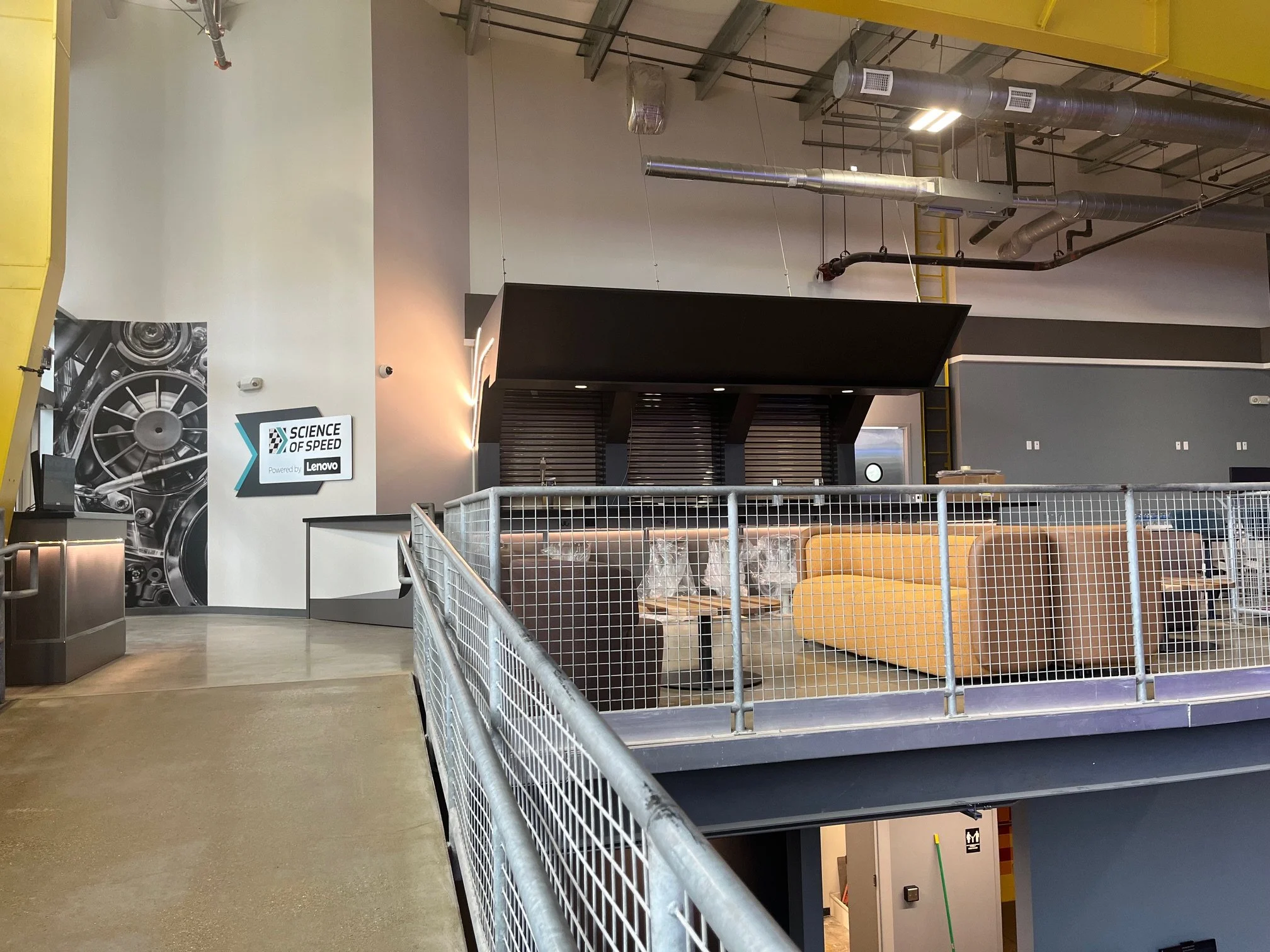 Interior of science center with industrial design, featuring exposed ductwork on the ceiling, a mezzanine with a metal railing, and a wall with a "Science of Speed" sign powered by Lenovo. There are some chairs and a counter below.