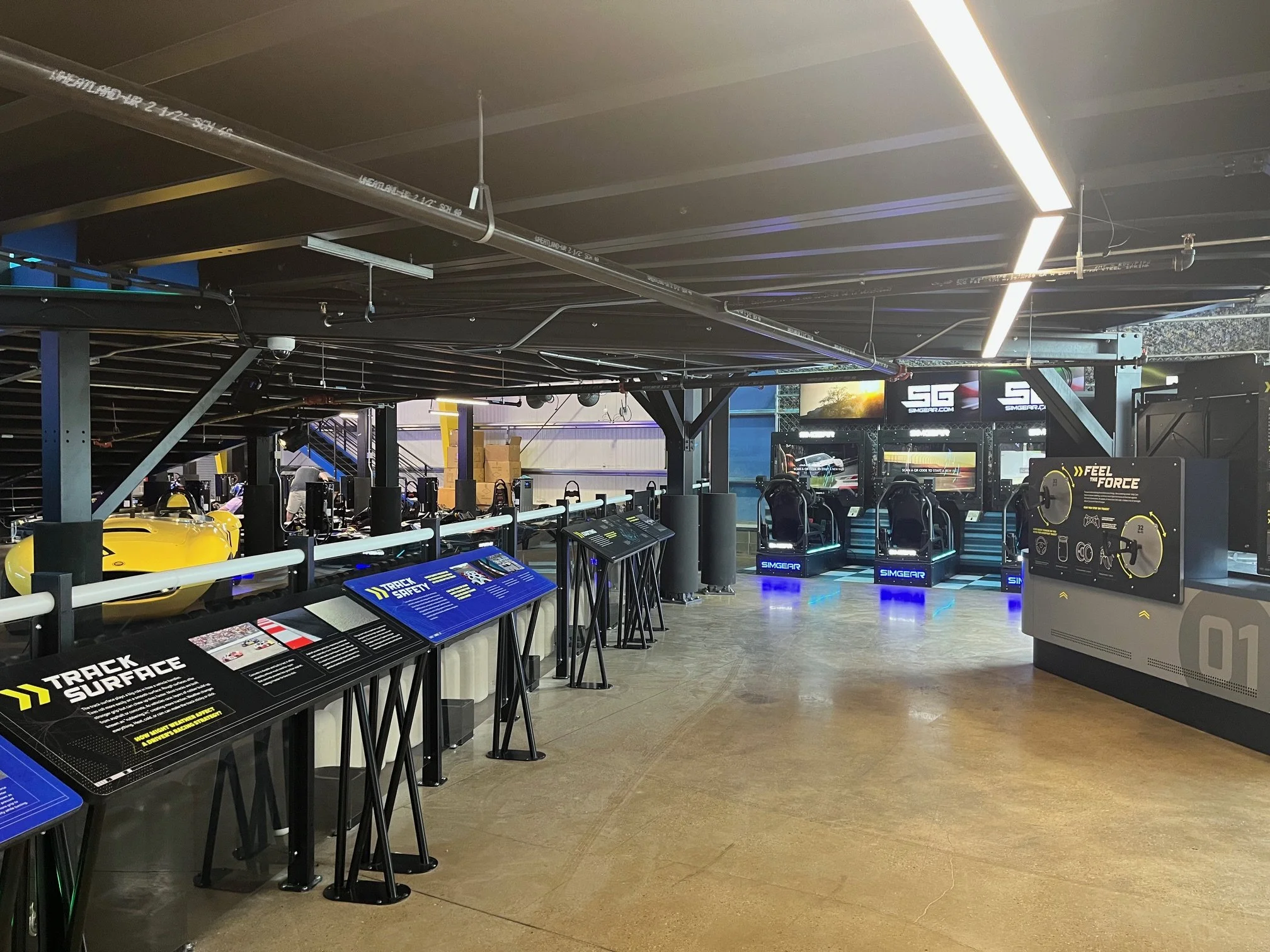 Indoor facility with workout equipment and informational display panels, featuring a large yellow sports car and electronic racing simulators.