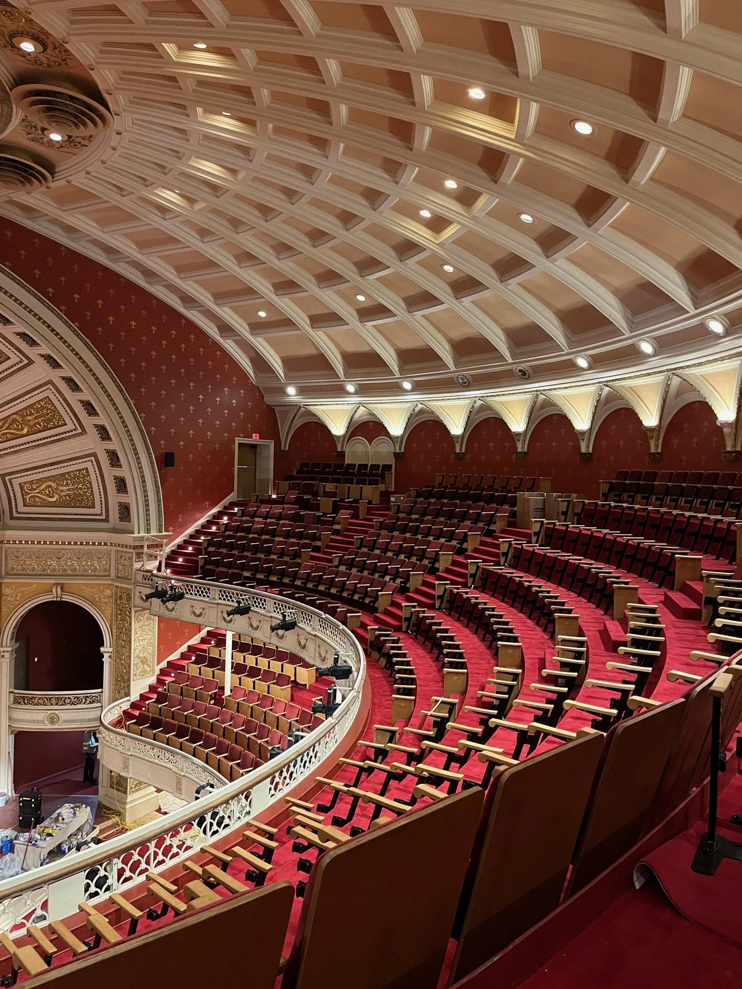 Interior view of a theater with red seats arranged in a curved pattern. The ceiling features ornate molding and recessed lighting. The walls are decorated with gold accents and small arches, with a balcony level with additional seating. There is a pe