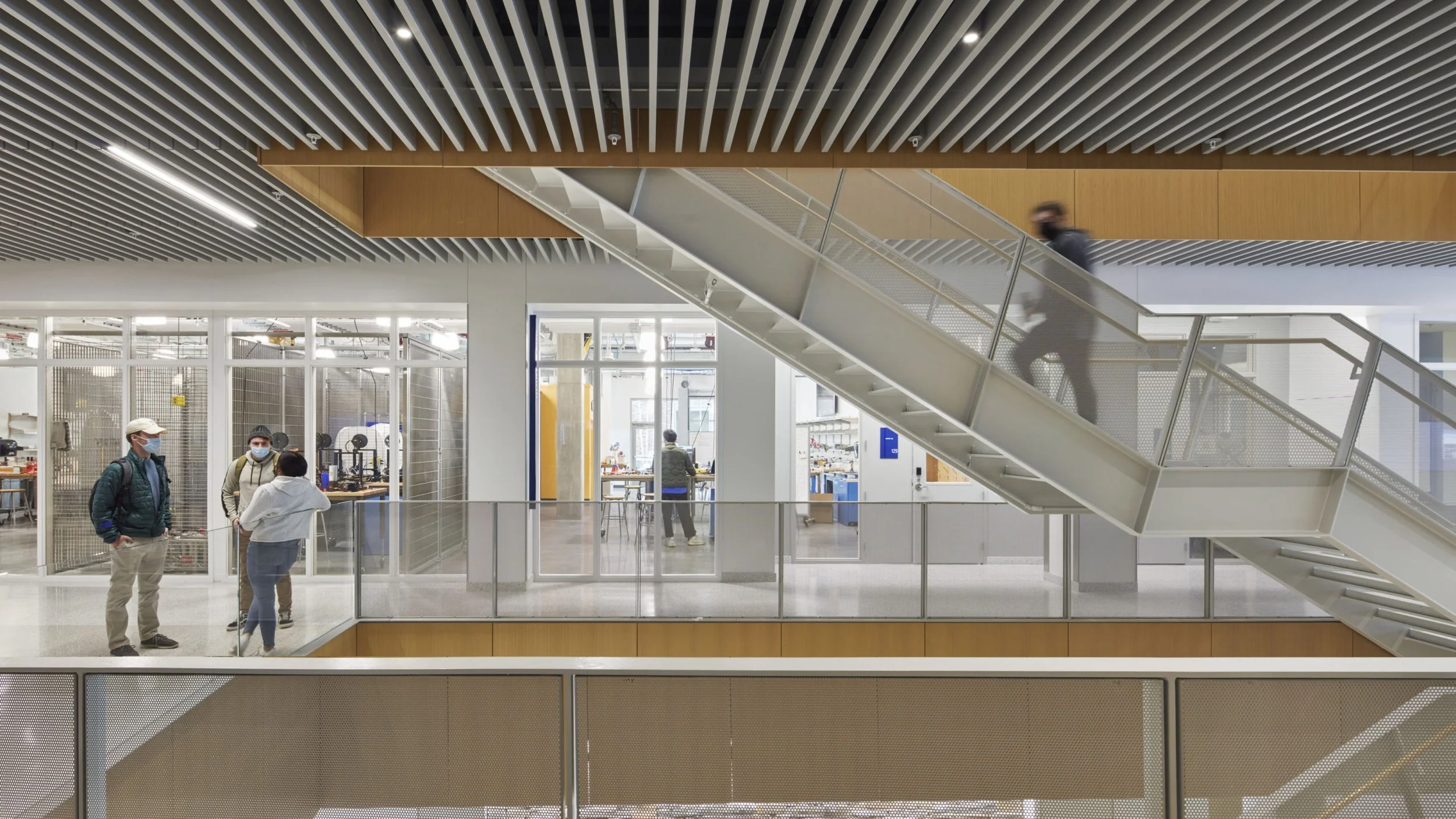 Multiple people wearing face masks inside a modern building with open workspaces, glass walls, and a staircase.