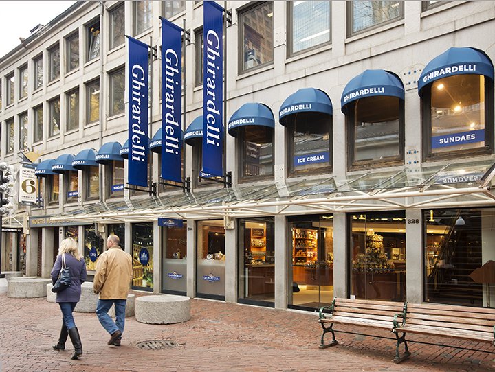 Ghirardelli ice cream and sundae shop on a city street with pedestrians walking by, blue flags and awnings displaying the Ghirardelli logo.