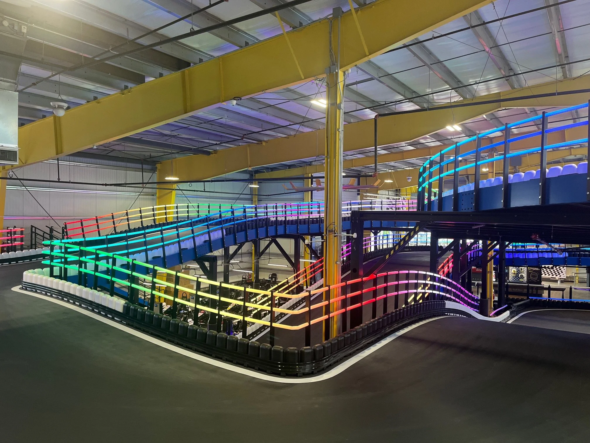 Indoor go-kart track with illuminated colorful neon lights, yellow overhead beams, black barriers, and a curved race path.