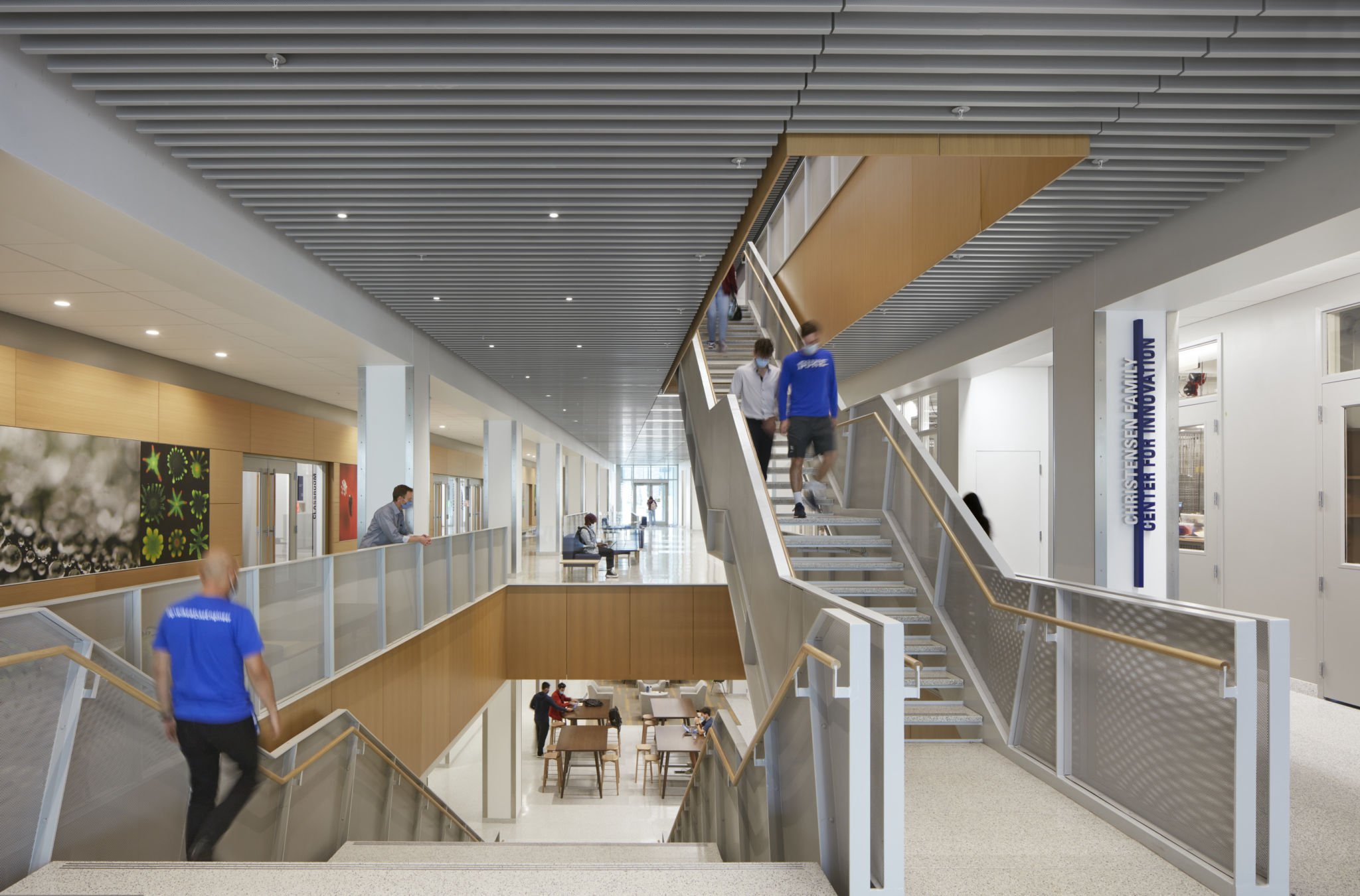 Interior of a modern building with stairs, walkways, and seating areas, people with masks, a sign for a university.