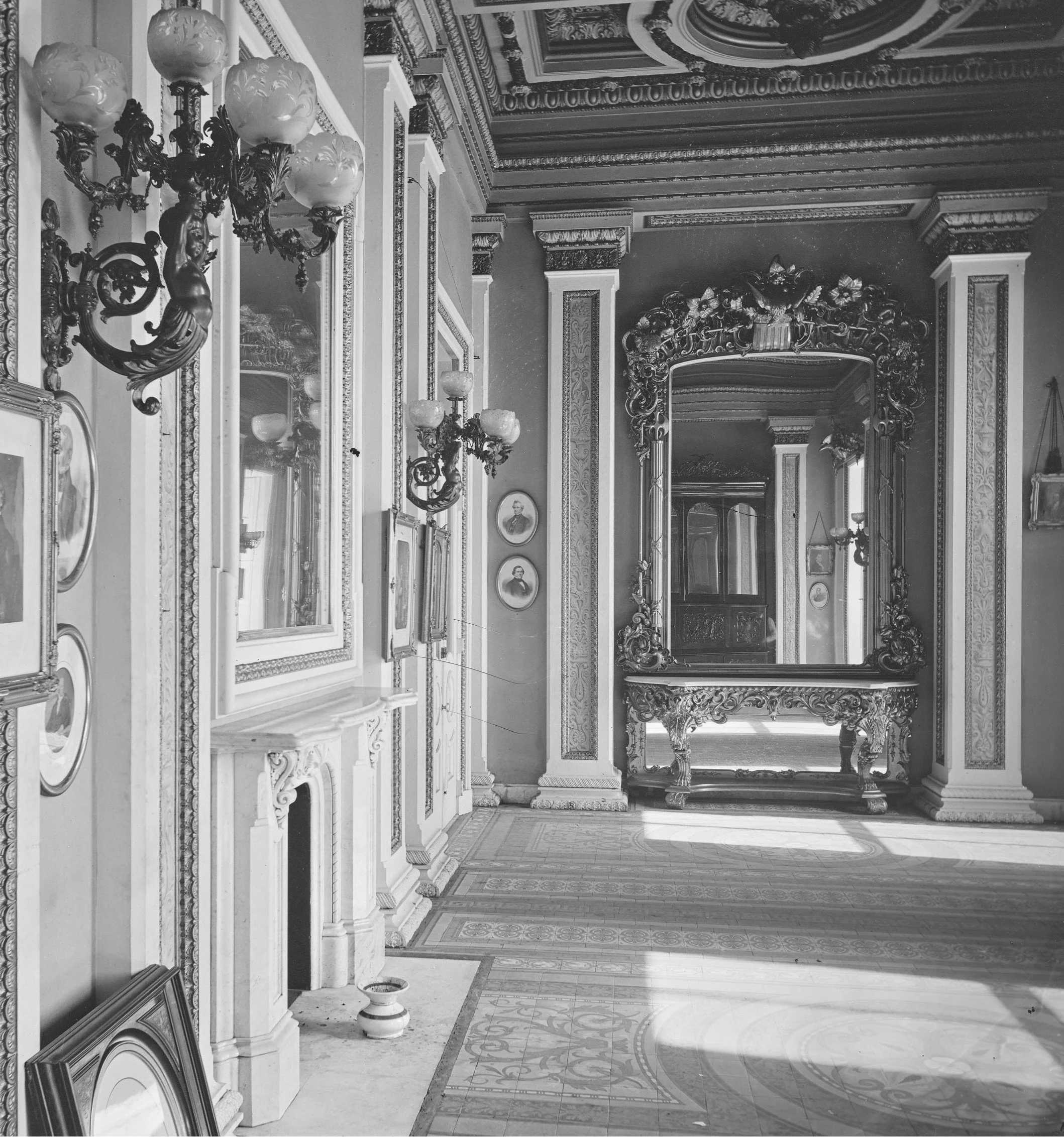 Ornate interior room with decorative moldings, elaborate mirrors, chandeliers, and framed photographs or paintings on the wall. The space features intricate woodwork and detailed wall and ceiling designs, suggesting a historic or luxurious setting.