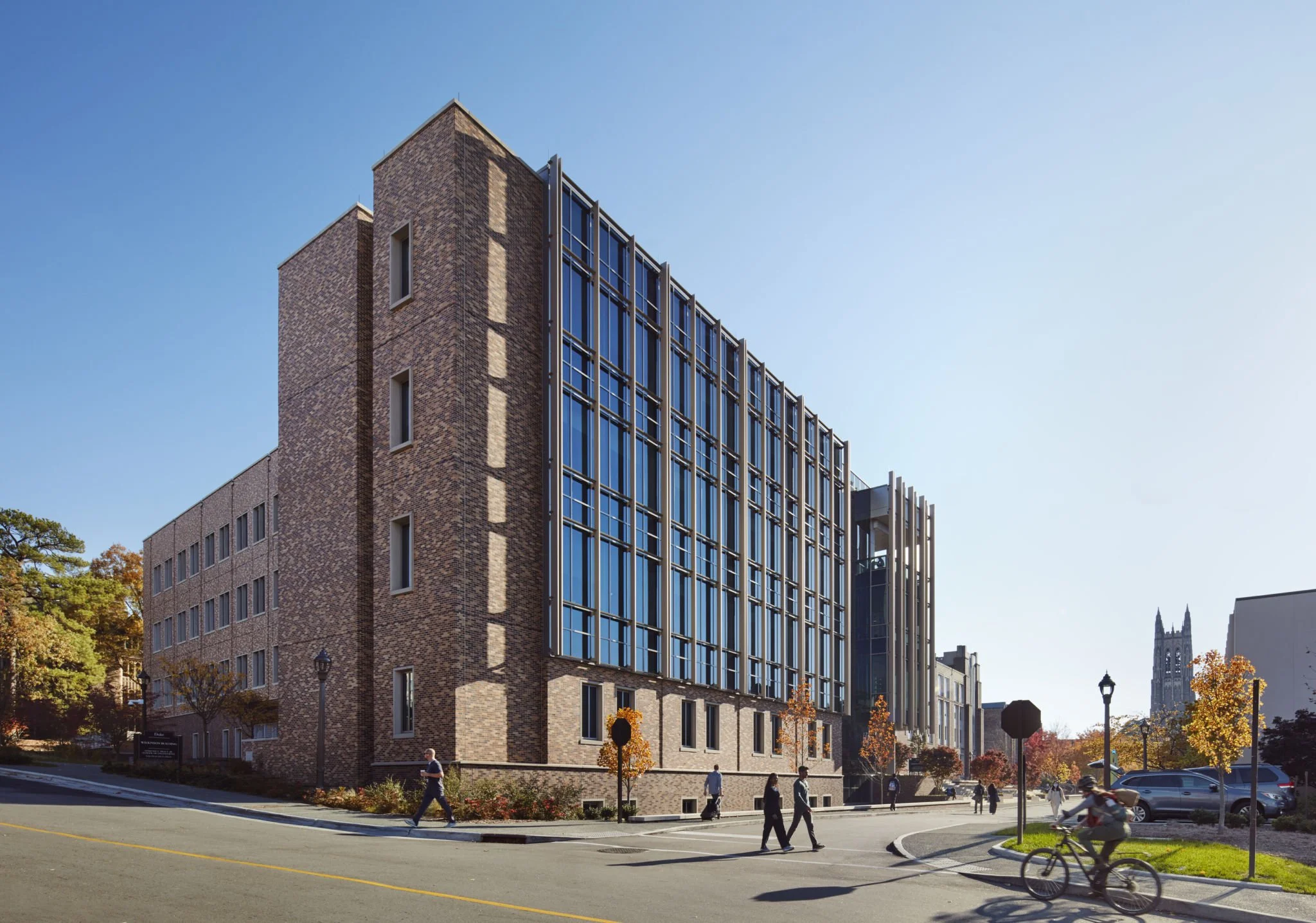 Modern brick and glass building on a city street with trees, people walking, and a cyclist.