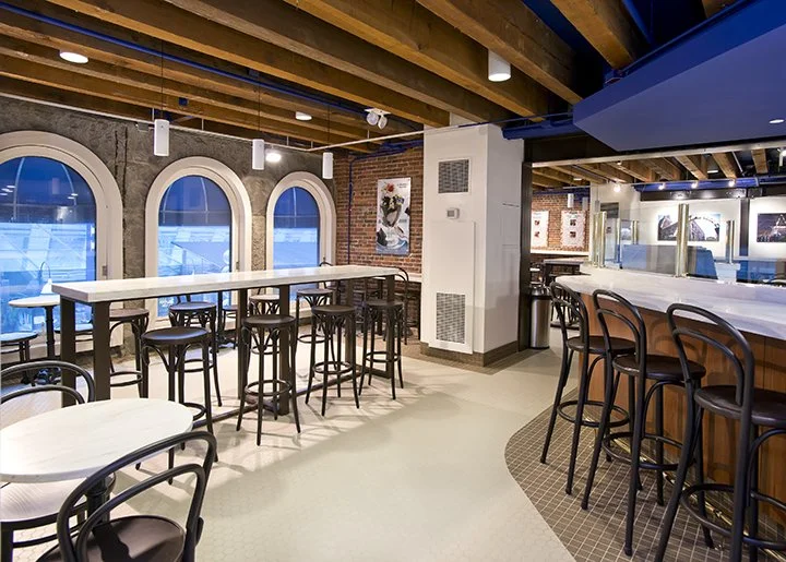 Interior of a modern restaurant or cafe with wooden beams, large arched windows, bar seating, and artwork on the brick walls.