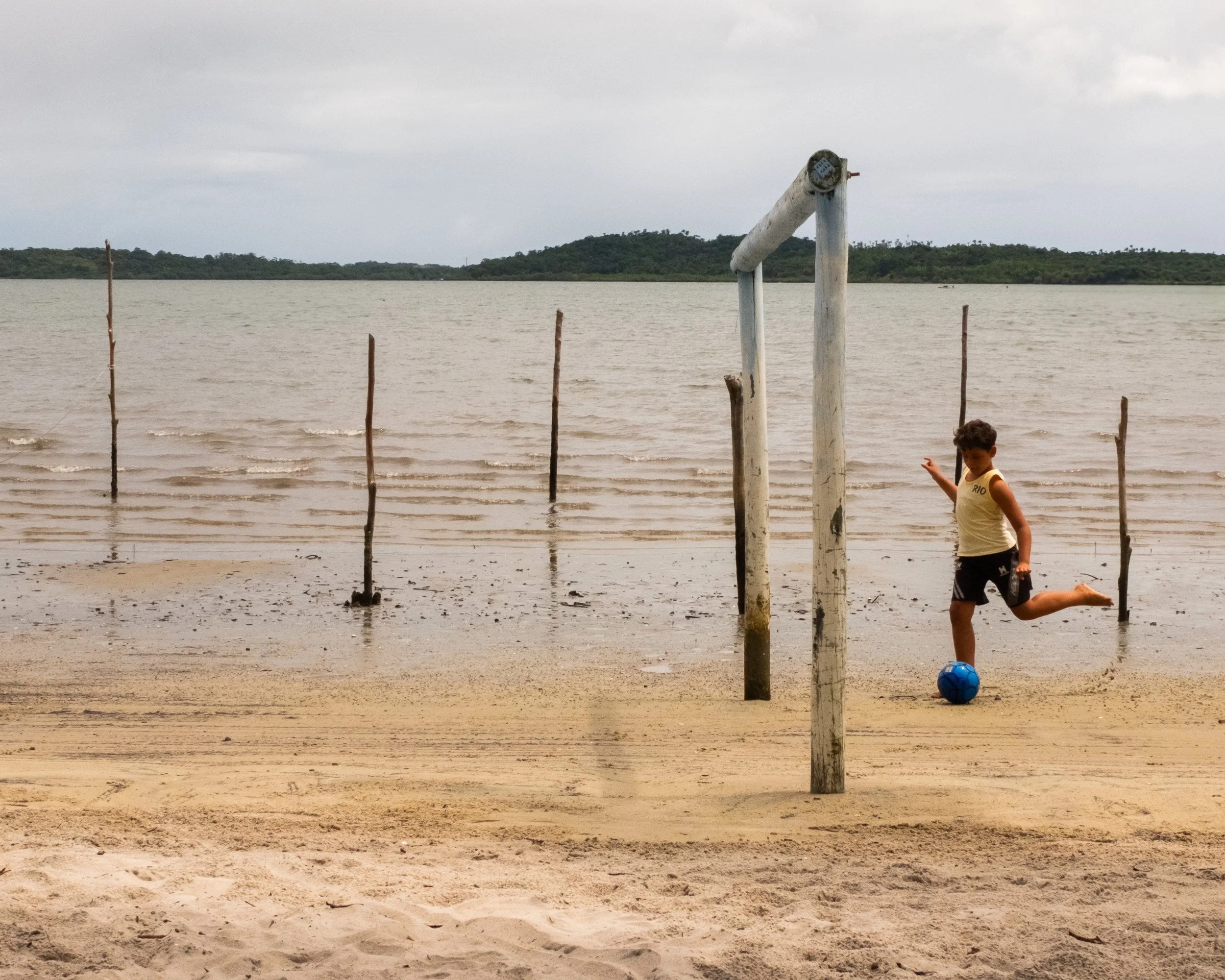 Criança jogando futebol na praia com uma bola azul, ao fundo há um mar tranquilo e um céu nublado.