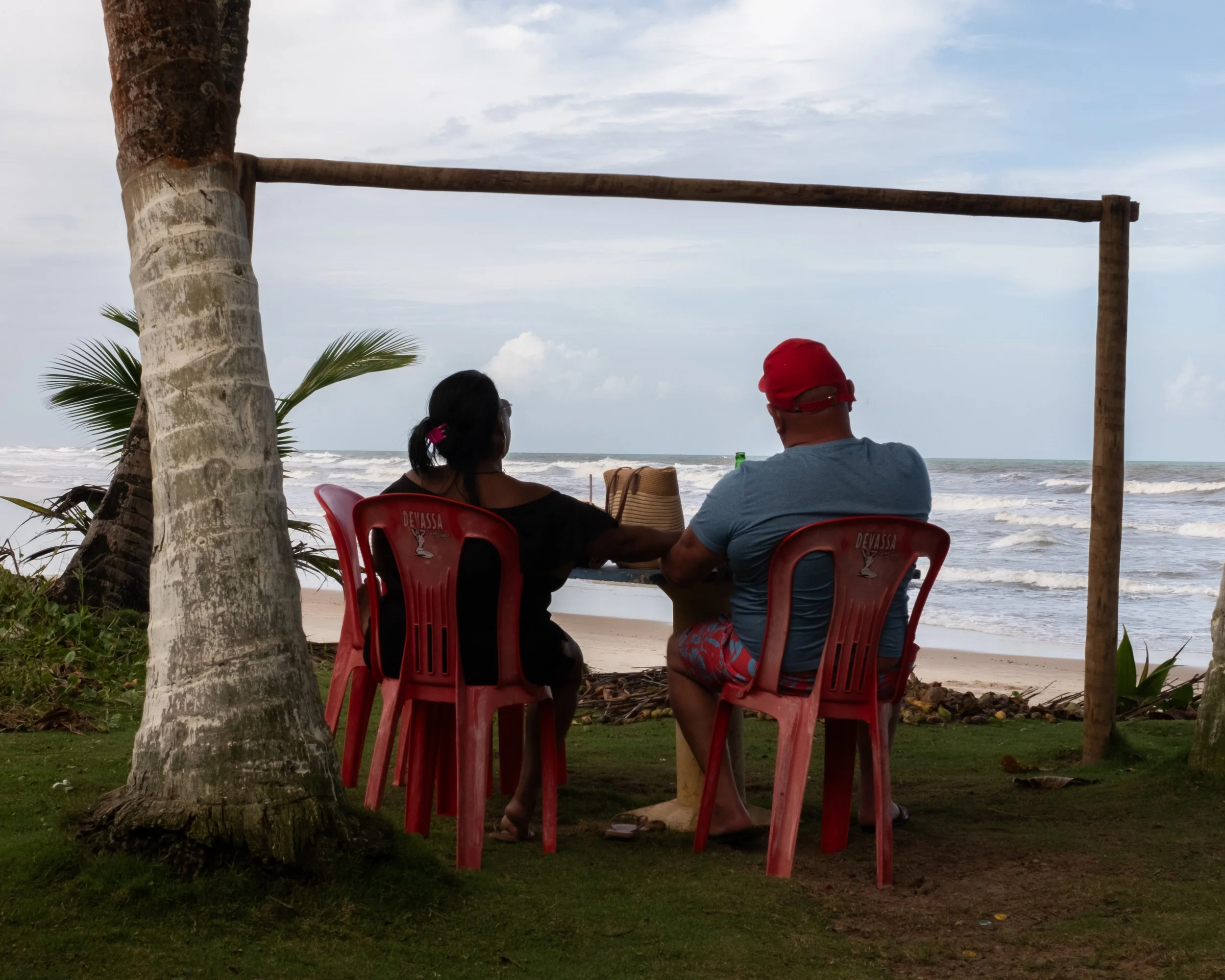 Homem e mulher sentados em cadeiras de plástico na praia, apreciando o pôr do sol ou o mar, sob uma estrutura de madeira e árvores ao redor.