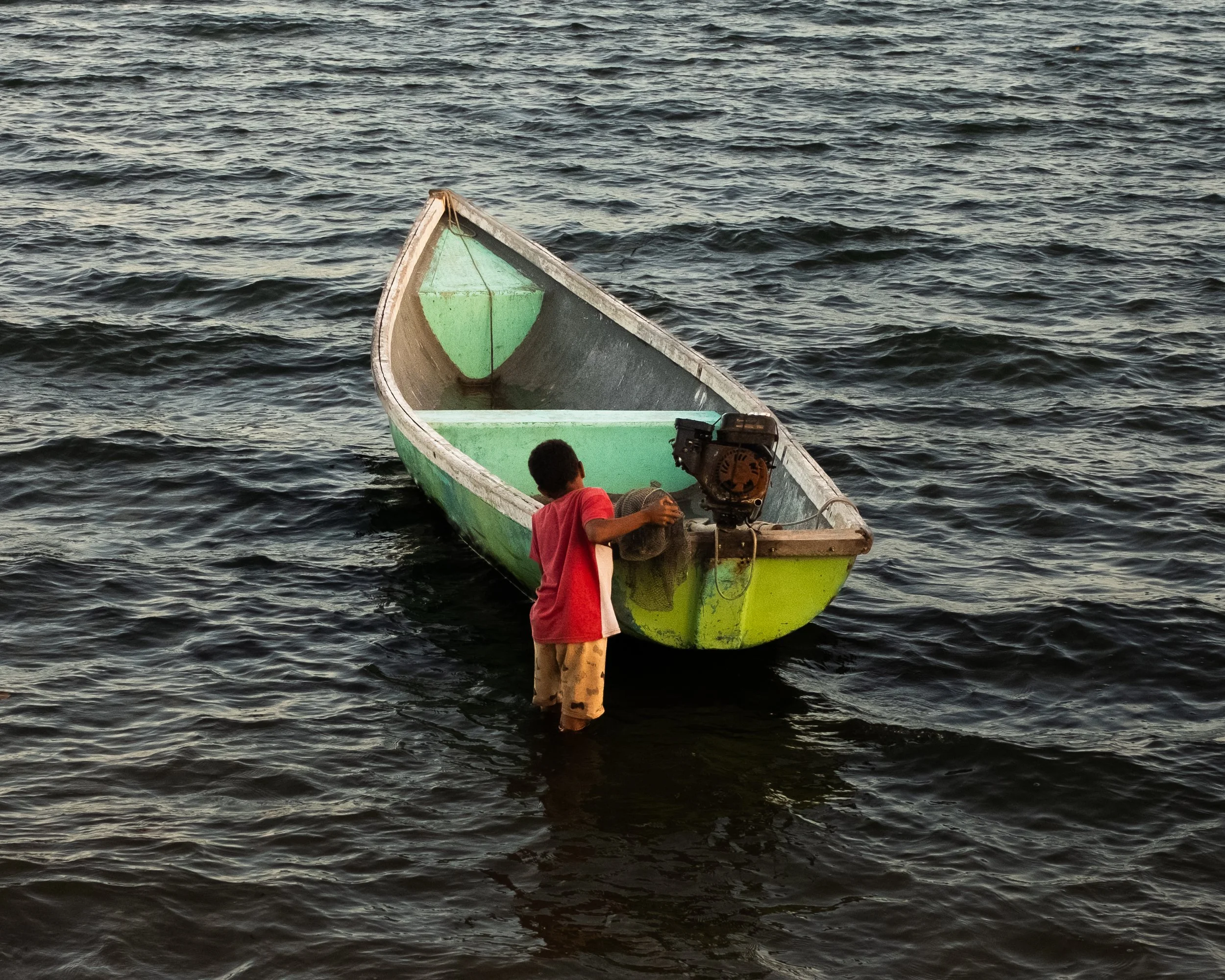 Criança de camiseta vermelha empurrando um barco verde ao mar.