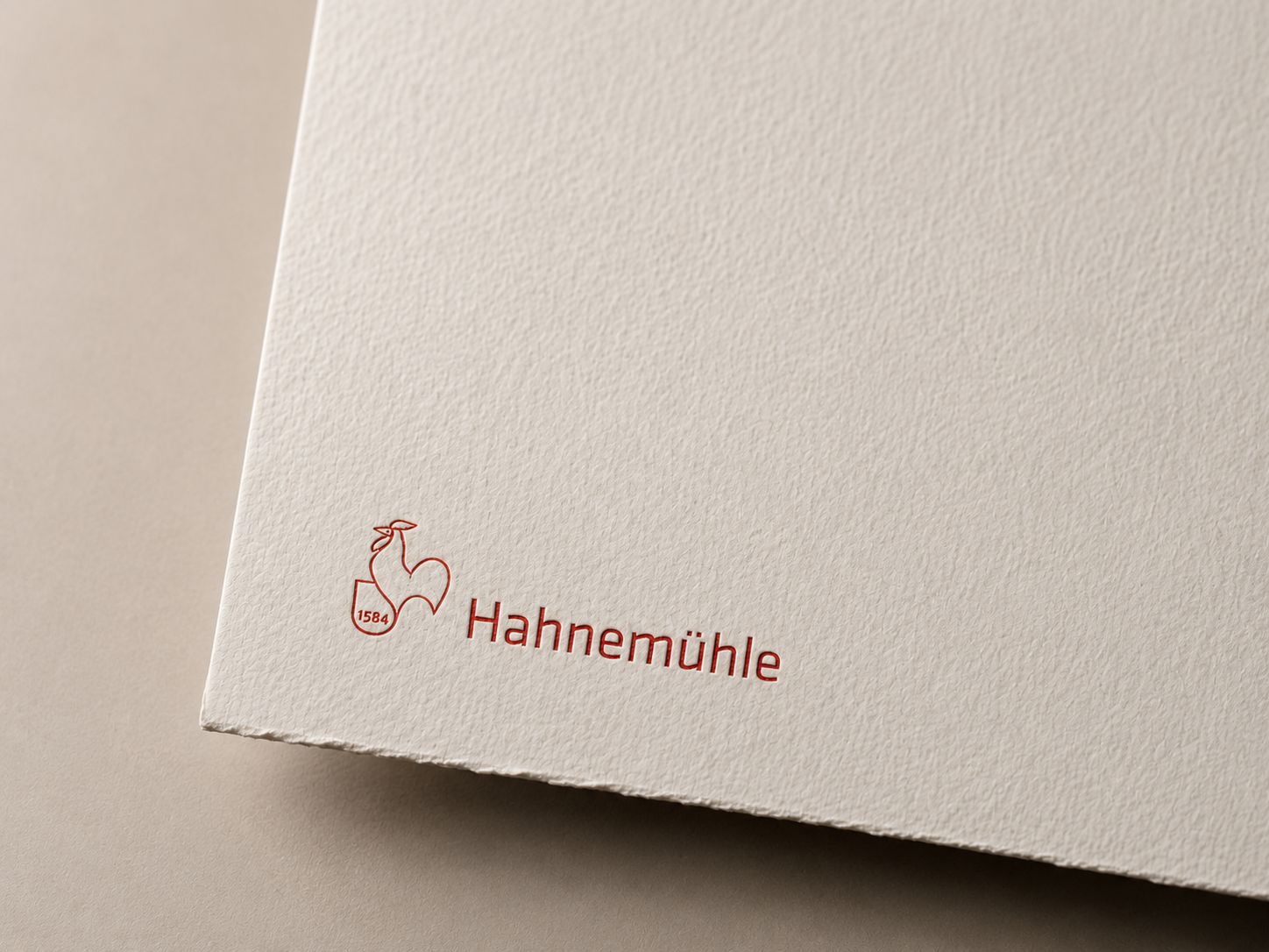 Logo da marca Hahnemühle com uma ilustração de uma cabra estilizada e o nome da marca em fonte vermelha, na parte inferior esquerda de uma embalagem de papel de alta qualidade.