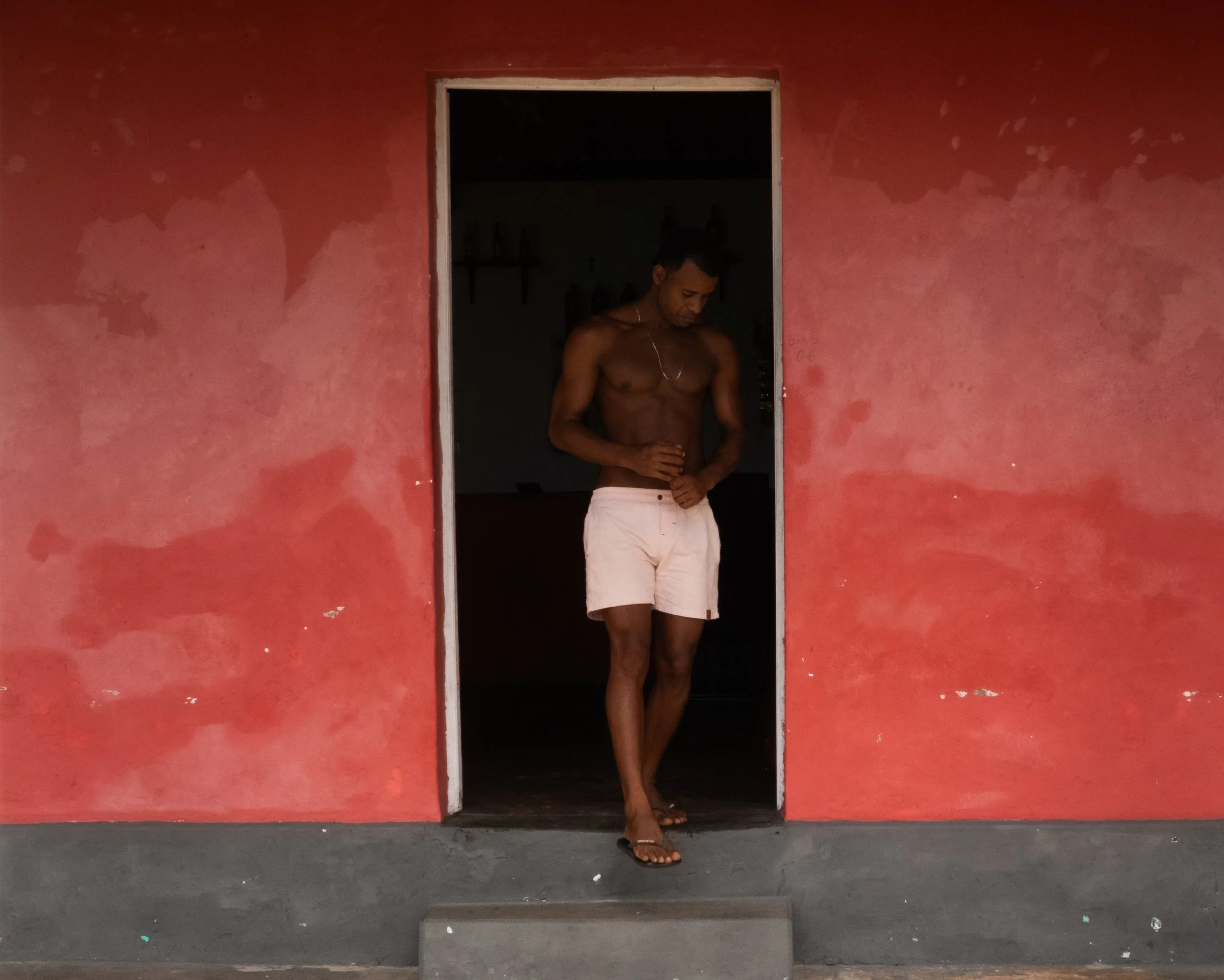 Homem jovem de olhos fechados, sem camisa e usando calça clara, de cabelos curtos, debruçado contra uma parede vermelha, na entrada de um cômodo escuro