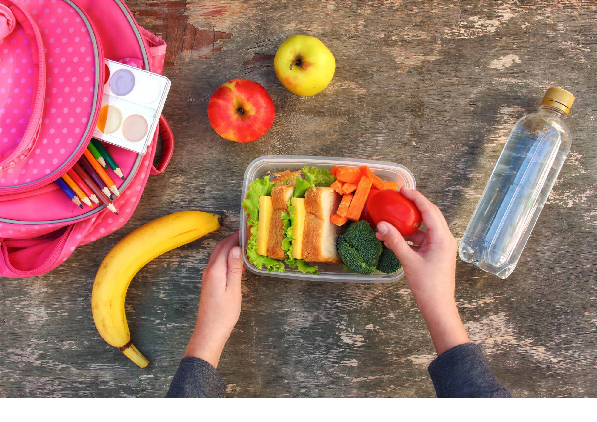 A person holding a lunch container with sandwiches, broccoli, and carrots. Nearby are apples, a banana, and a water bottle on a wooden table, with a pink polka-dotted backpack.