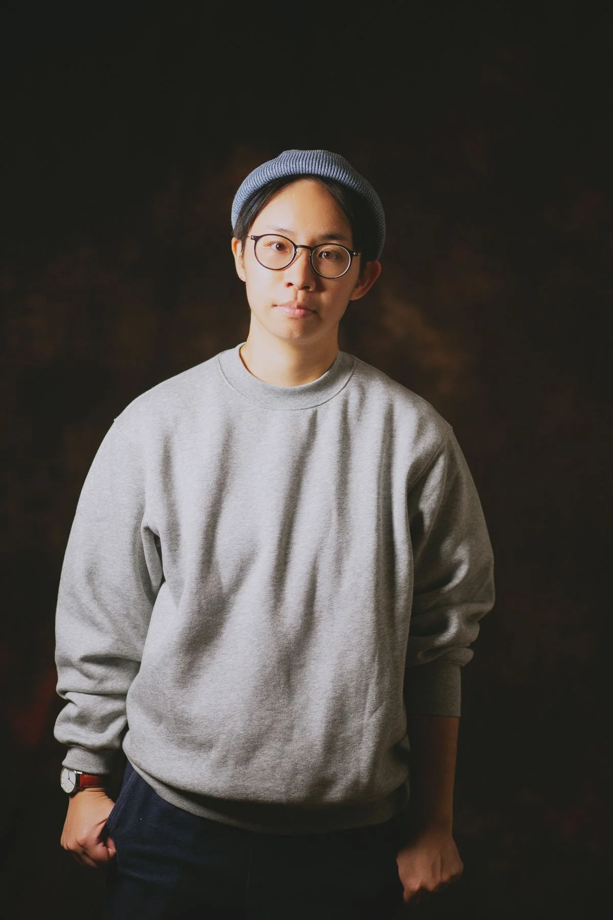 A young person wearing glasses, a gray sweatshirt, a watch, and a gray beanie hat standing against a dark background, with one hand in their pocket.