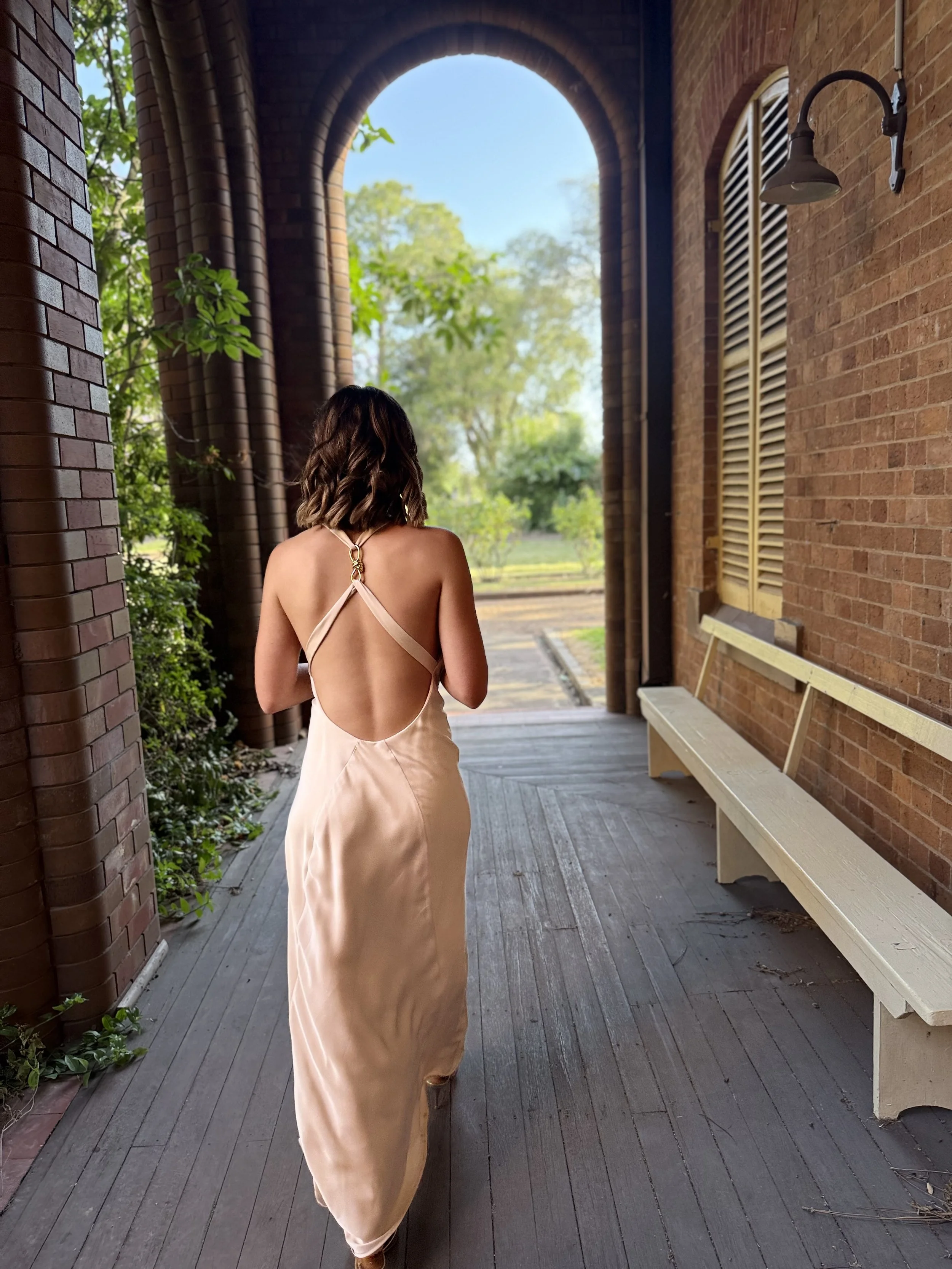 A woman wearing a backless, silky, beige dress walking on a wooden deck, with brick walls on either side and a bench on the right. Green trees are visible through an arched opening at the end of the porch, and there is a vintage wall-mounted outdoor light on the right wall.