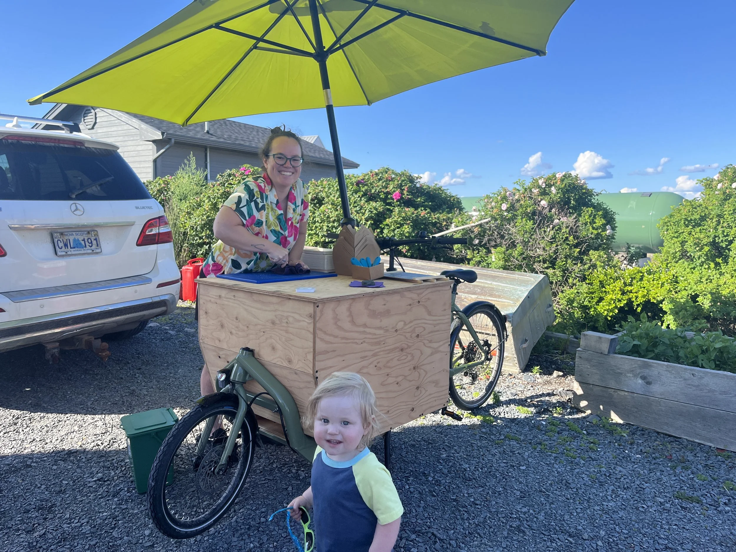 A smiling woman outdoors shucking oysters behind a wooden cart attached to a bicycle, with a large yellow-green umbrella attached, and a small child running in the foreground.