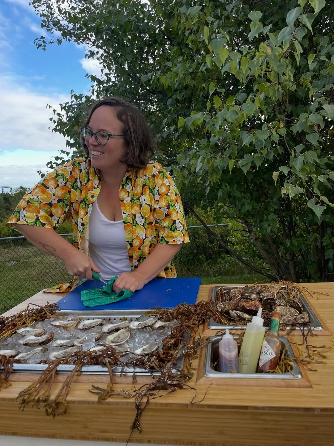 Woman in a yellow tropical shirt with glasses, shucking oysters on a blue cutting board at an outdoor table, surrounded by seafood and condiments.