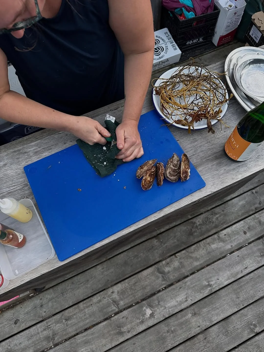 Person preparing oysters on a blue cutting board with oyster shells, a plate of seaweed, and condiments on a rustic outdoor kitchen counter.