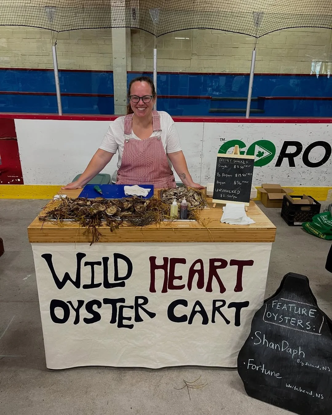 A woman is smiling at a seafood stand labeled "Wild Heart Oyster Cart" at an indoor market. The stand displays fresh oysters on a wooden table, with a chalkboard menu indicating prices for shucked and unshucked oysters. The woman wears glasses and a red and white striped apron