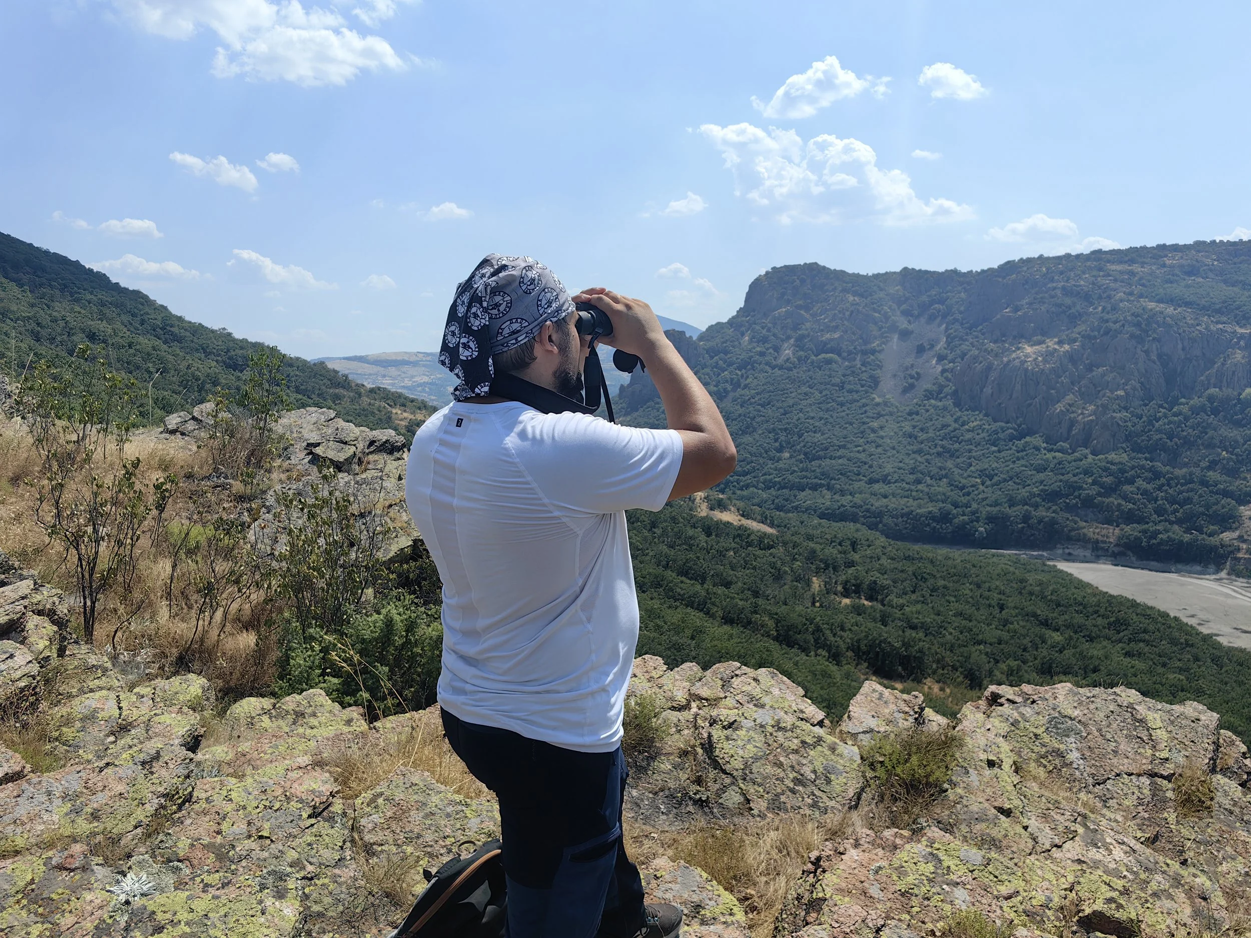A man wearing a bandana, white t-shirt, and black shorts is standing on rocky terrain, looking through binoculars at a scenic view of green hills, cliffs, and a river under a partly cloudy sky.