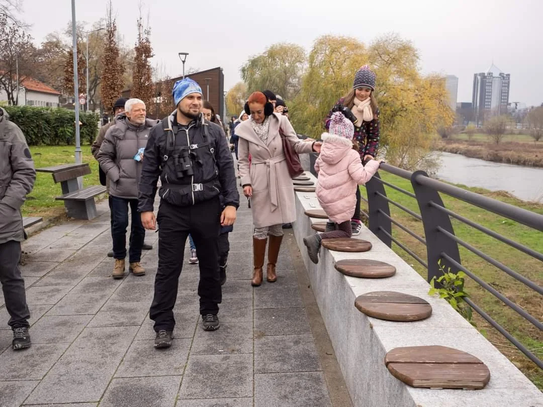 People walking on a paved pathway beside a river on a cloudy day. Two children are climbing onto a concrete barrier with wooden circles, while several adults walk nearby. The background features trees with autumn foliage and a few tall buildings.