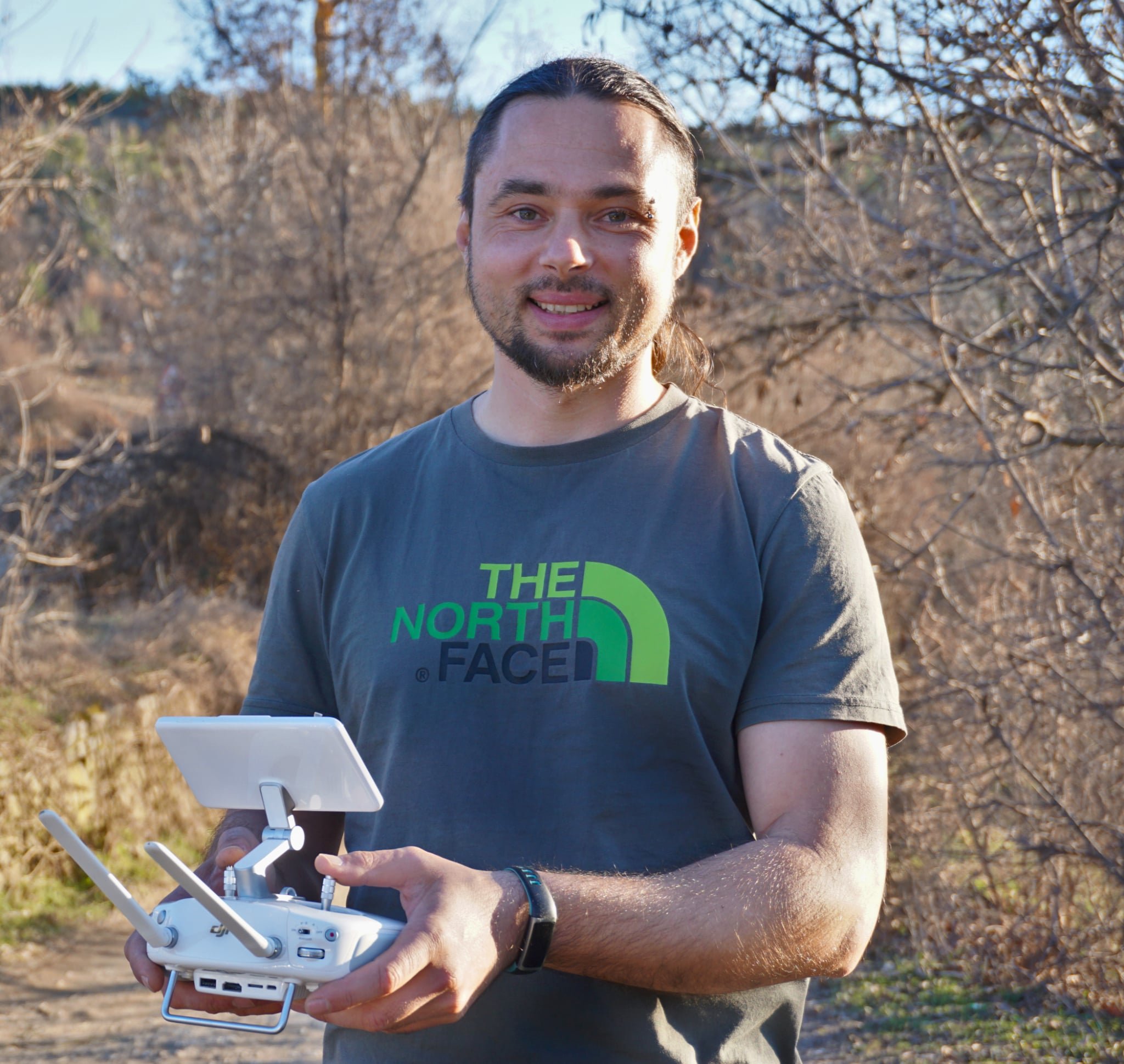 A man wearing a gray T-shirt with The North Face logo, holding a drone controller outdoors in a wooded area with leafless trees.