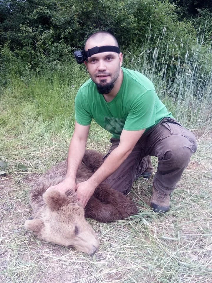 A man kneeling on the grass, holding a bear cub in a natural outdoor setting with green foliage in the background.