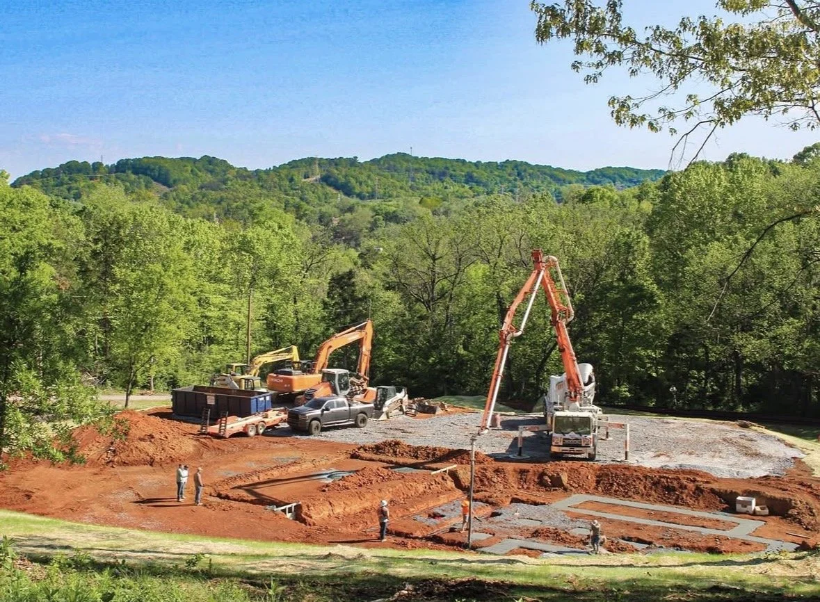 Construction site with excavators and workers preparing foundation in a green, wooded area with hills in the background.
