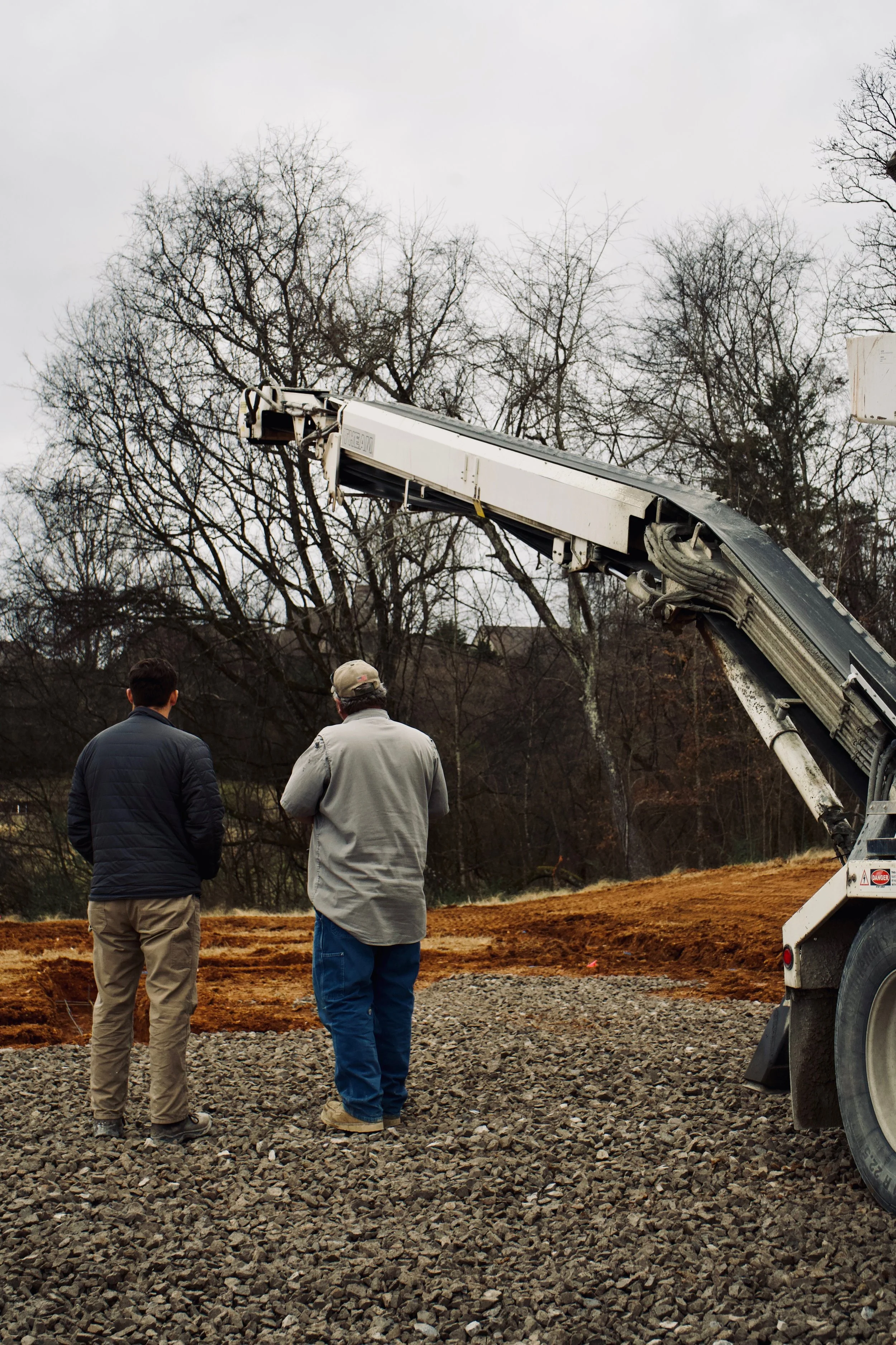 Two men standing outdoors on rocky ground, observing a large construction crane lifting a load, with bare trees and cloudy sky in the background.