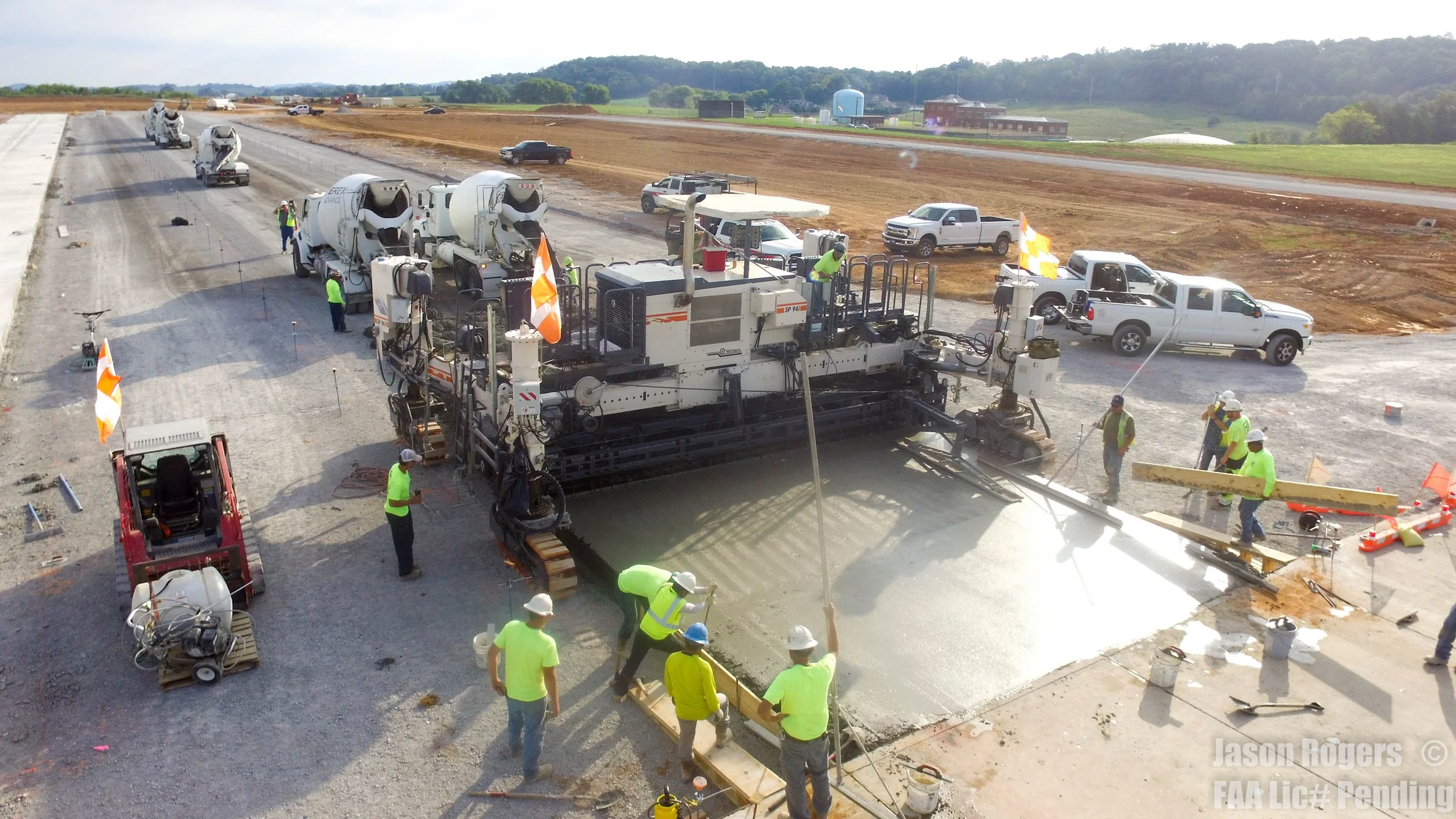 Construction workers pouring concrete on a road at a construction site, with trucks, construction equipment, and vehicles surrounding the area.