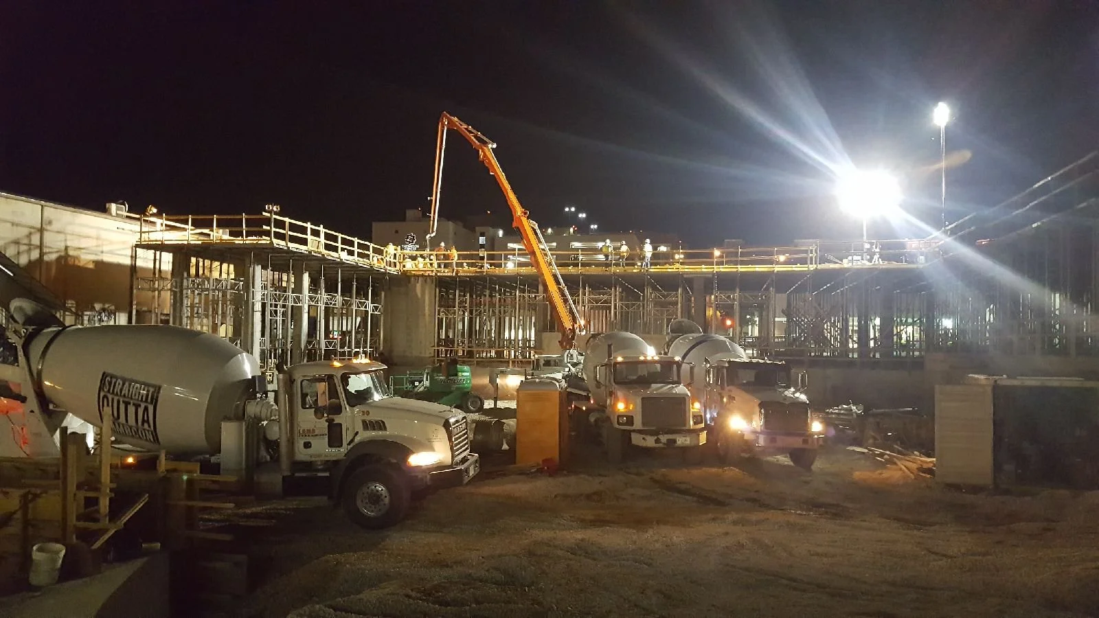 Nighttime construction site with concrete mixers, a crane, and workers on a scaffold.