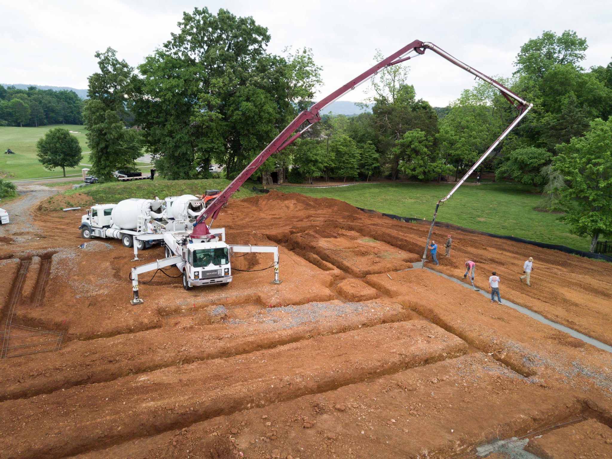 Construction site with excavated area, concrete mixer truck, and workers, surrounded by green trees and grassy fields.