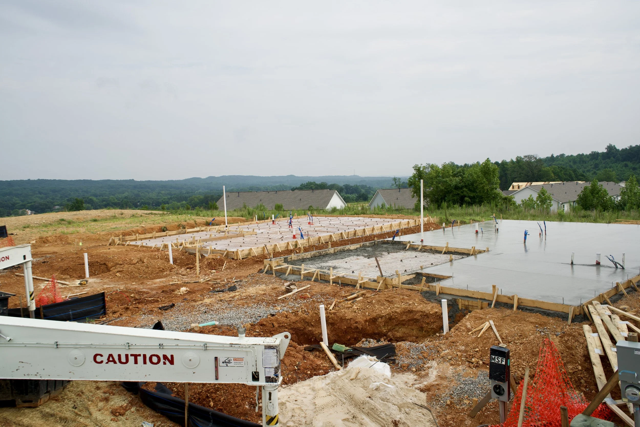 Construction site with concrete foundation and wooden framework, caution signs and pipes protruding, residential houses and green hills in the background.