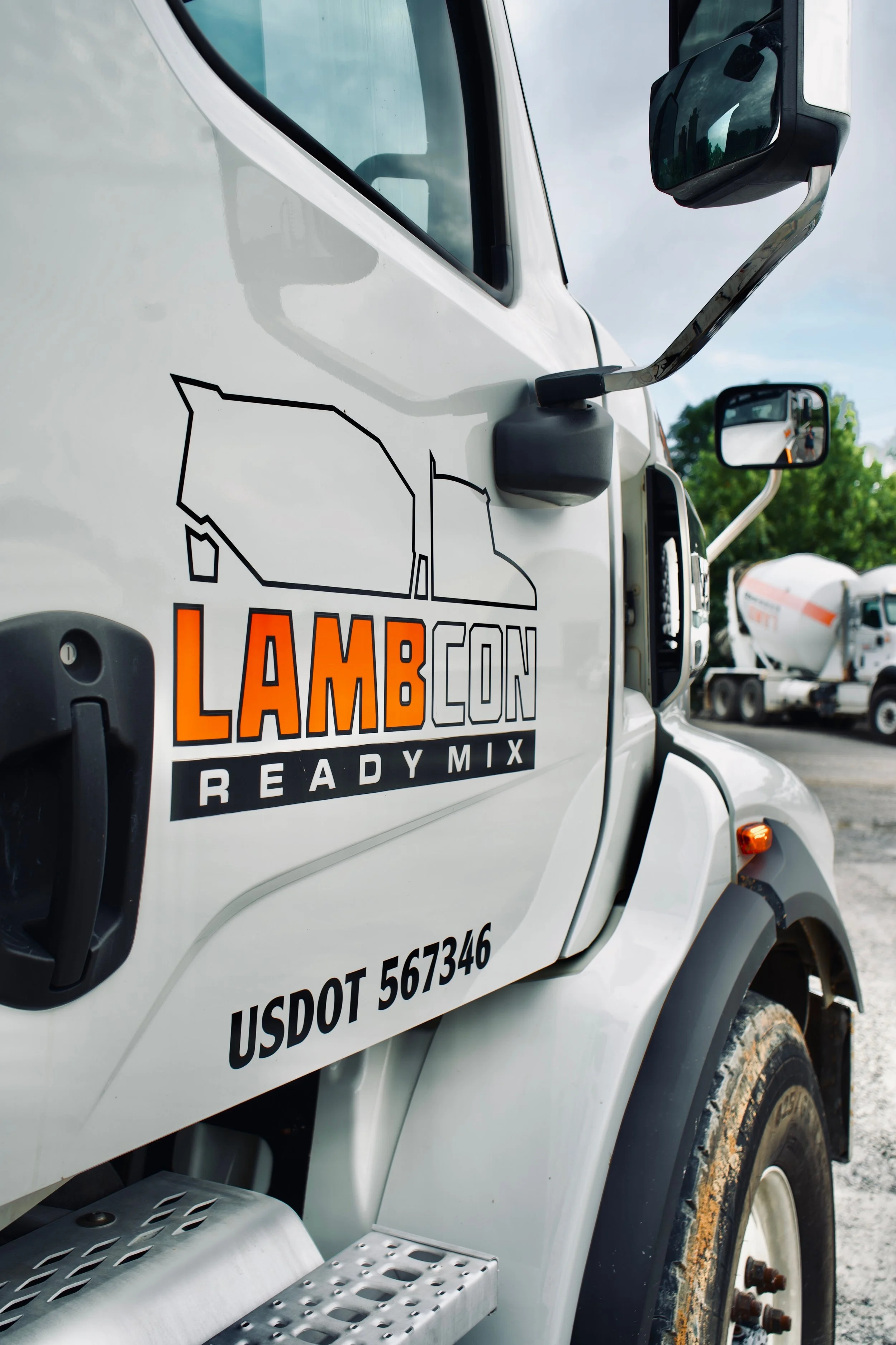 A white livestock truck with the logo 'LAMBCON READY MIX' on the side, showing the cab with side mirror, fuel tank, and part of the tire.